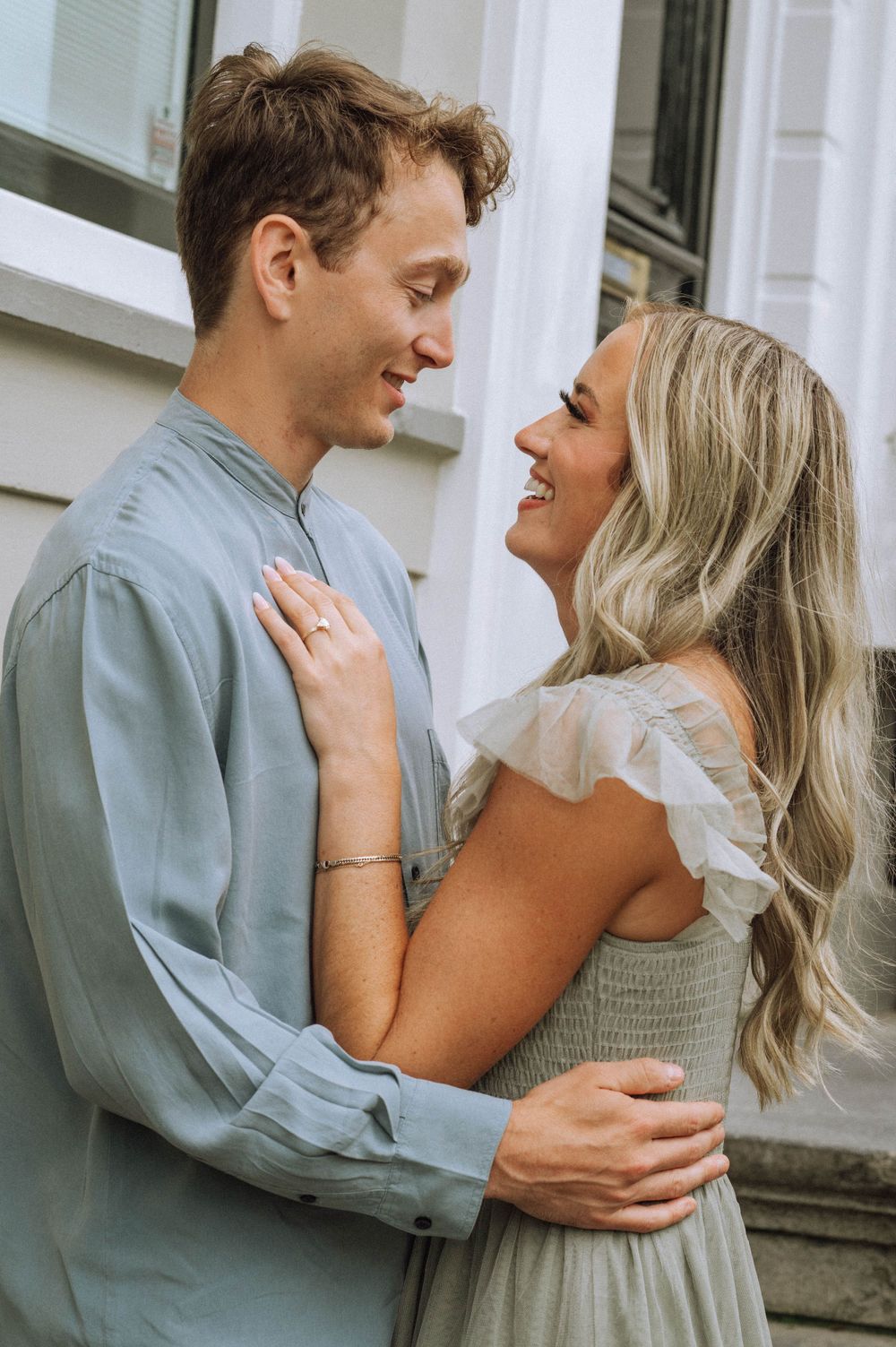 A couple shares an intimate embrace against a white building with green shutters.