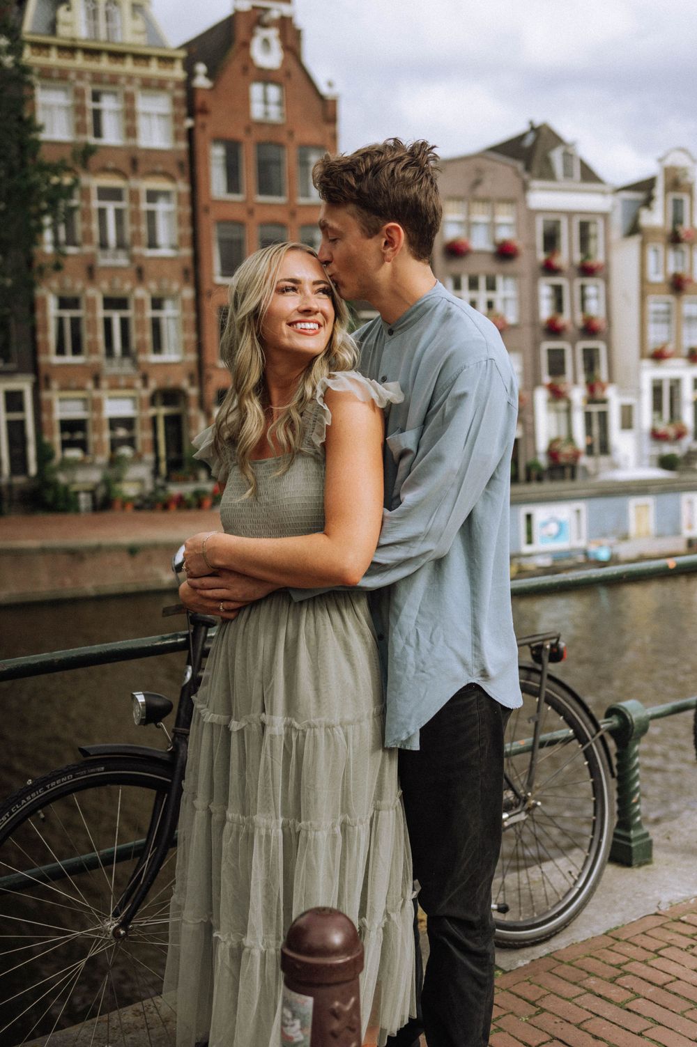 Romantic couple shares tender moments along a canal in Amsterdam with historic buildings in the background.