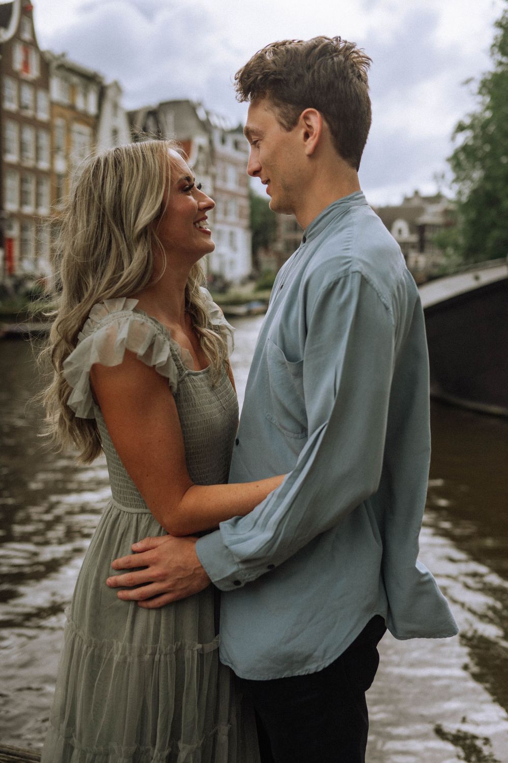 Couple embraces by a scenic canal in Amsterdam wearing coordinating sage green and blue outfits.