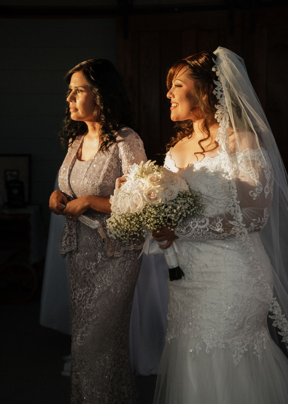 Two people in formal attire stand together in a dimly lit room, one wearing a lace gown and veil.