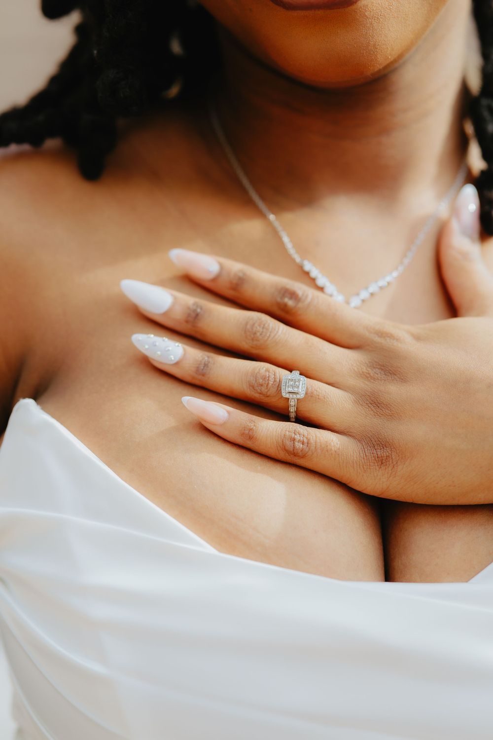 Close-up of hands with white manicured nails wearing an elegant diamond engagement ring and delicate necklace against white fabric.