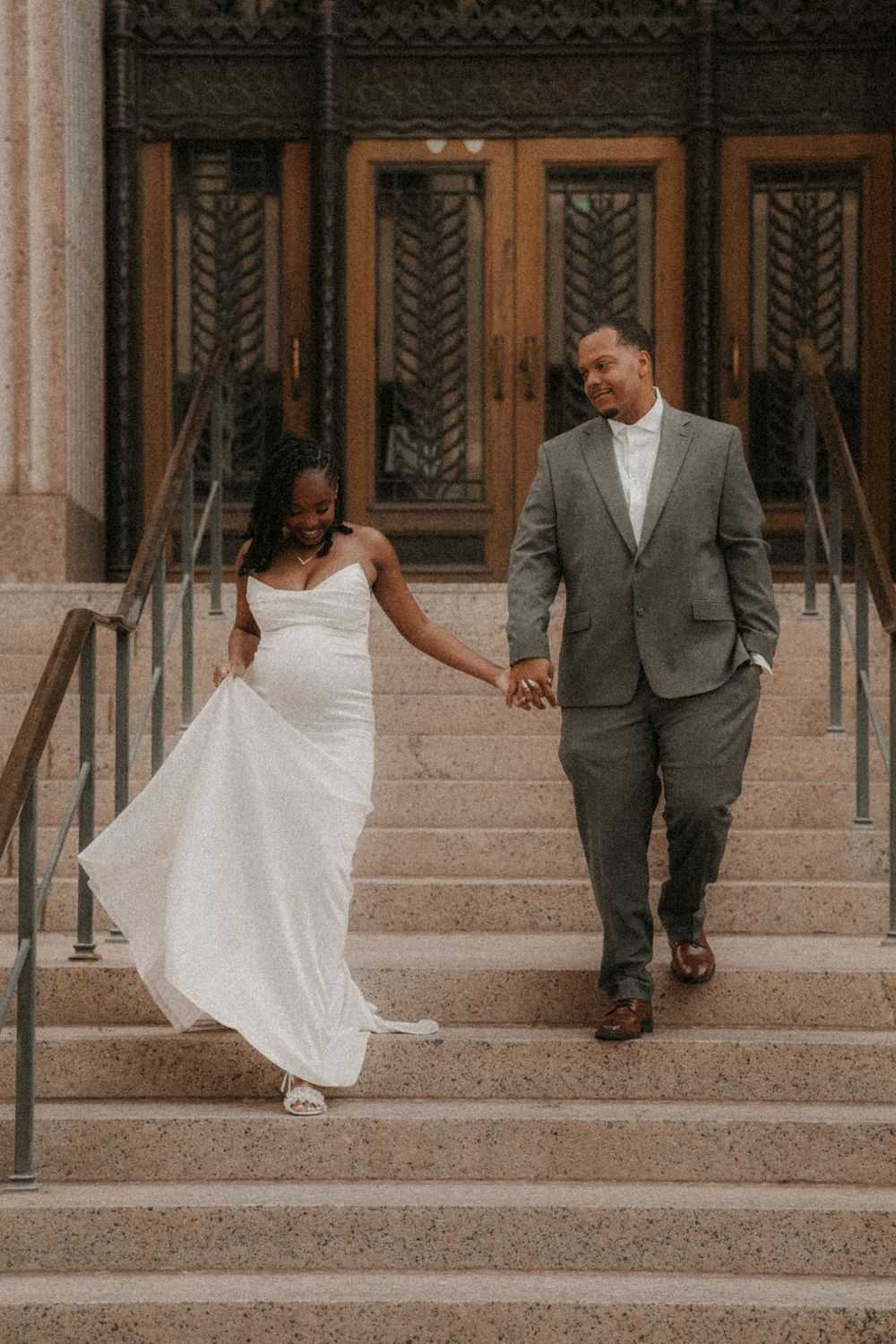 A couple descends stone stairs together, with the bride in a flowing white dress and groom in a gray suit.