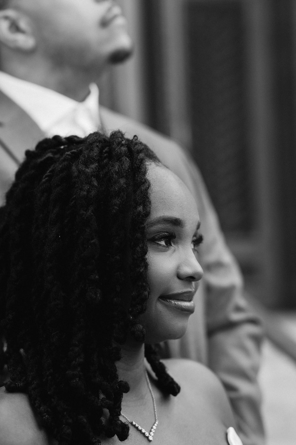 A profile view of a person with curly black hair wearing a necklace smiling in a black and white photograph.