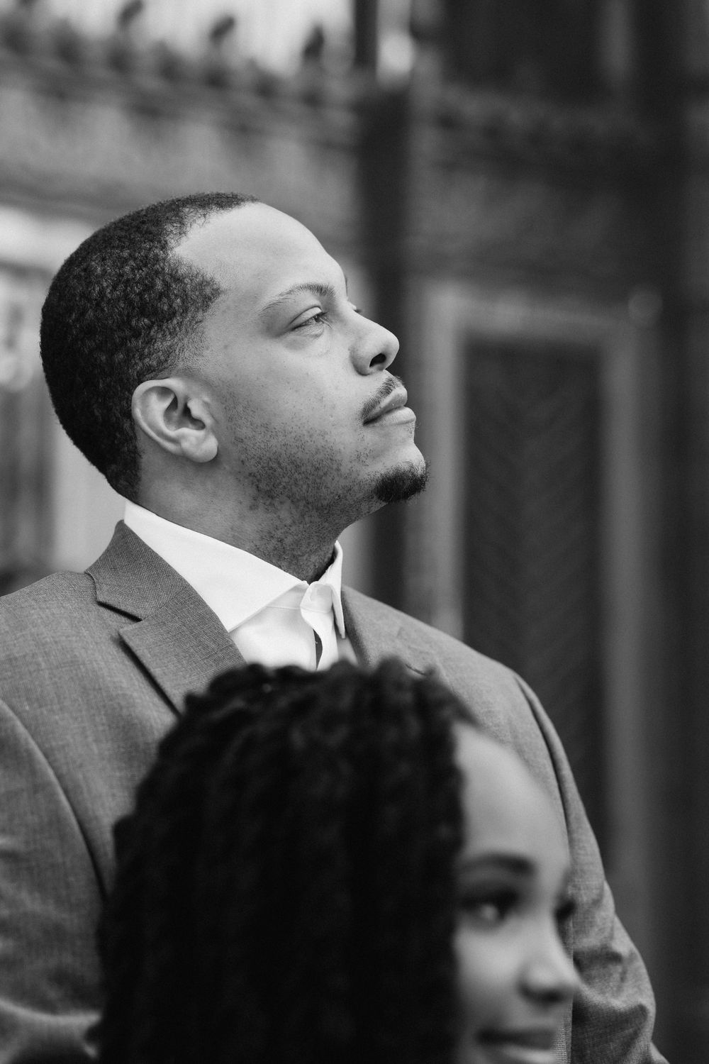 A black and white candid portrait of a person in a suit looking upward with a contemplative expression.