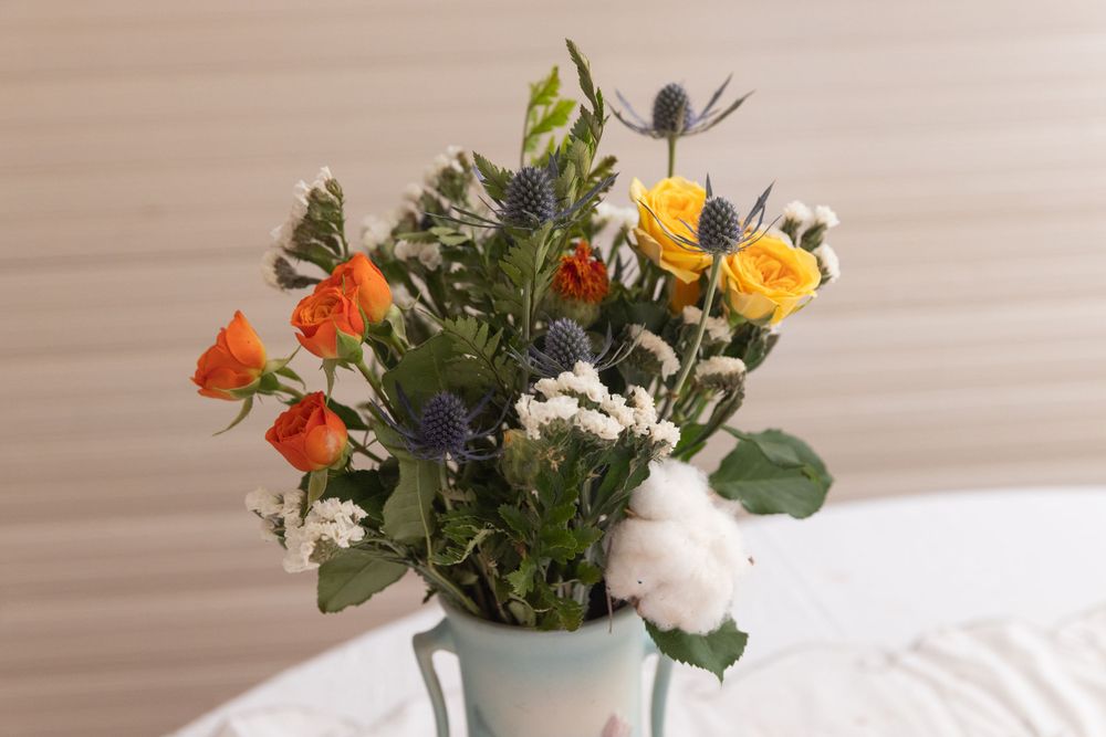 A bright bouquet of orange, yellow, and white flowers arranged in a light blue vase against a beige wall.
