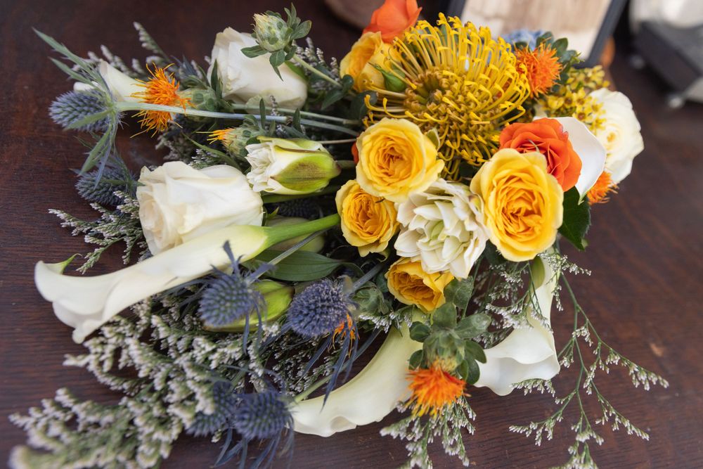 A vibrant wedding bouquet featuring yellow roses, white calla lilies, orange flowers and blue thistle on a dark wooden surface.