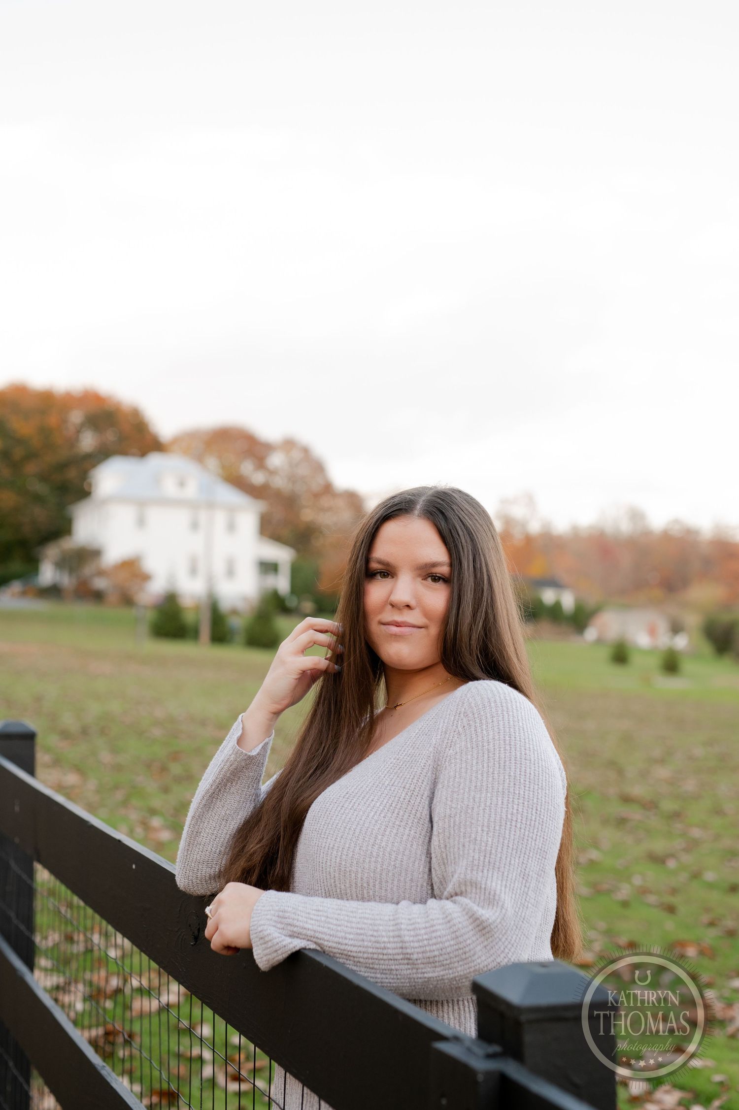 Casual fall portrait by wooden fence with white house and autumn trees in background.