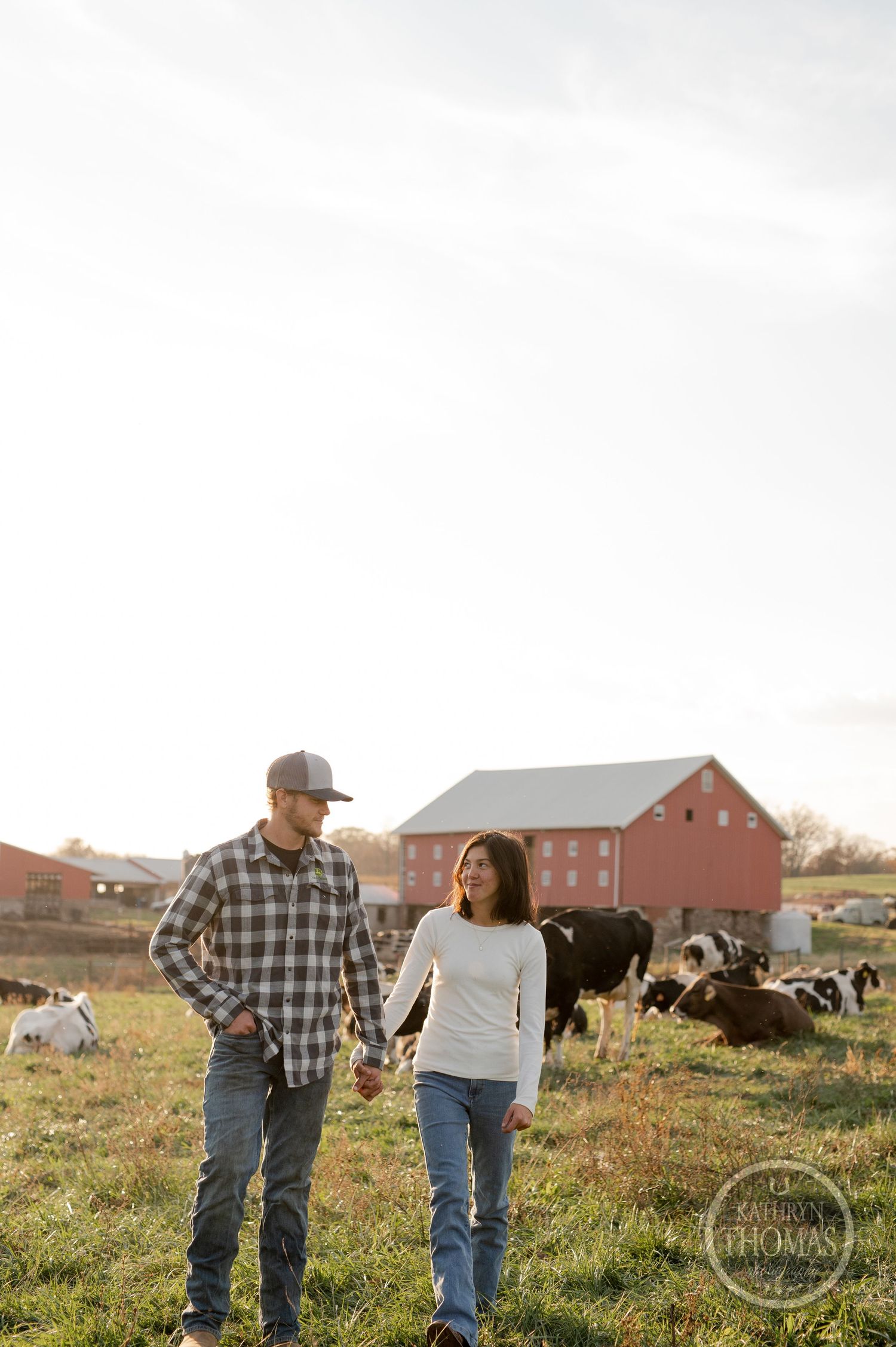 A couple walks together on their farm property with grazing cattle and a red barn in the background.