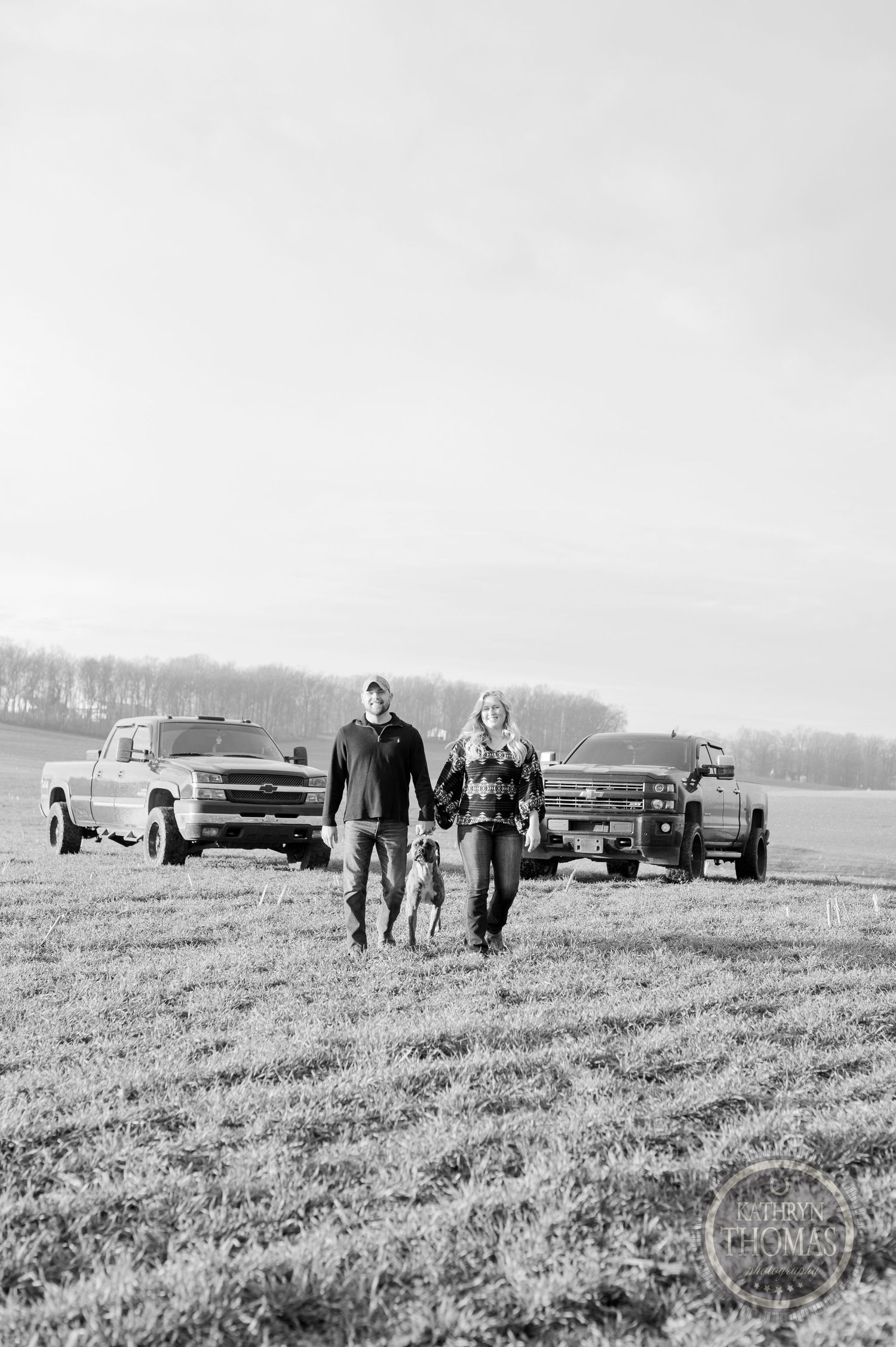 A black and white photo of a couple walking with their dog in a field with trucks in the background.
