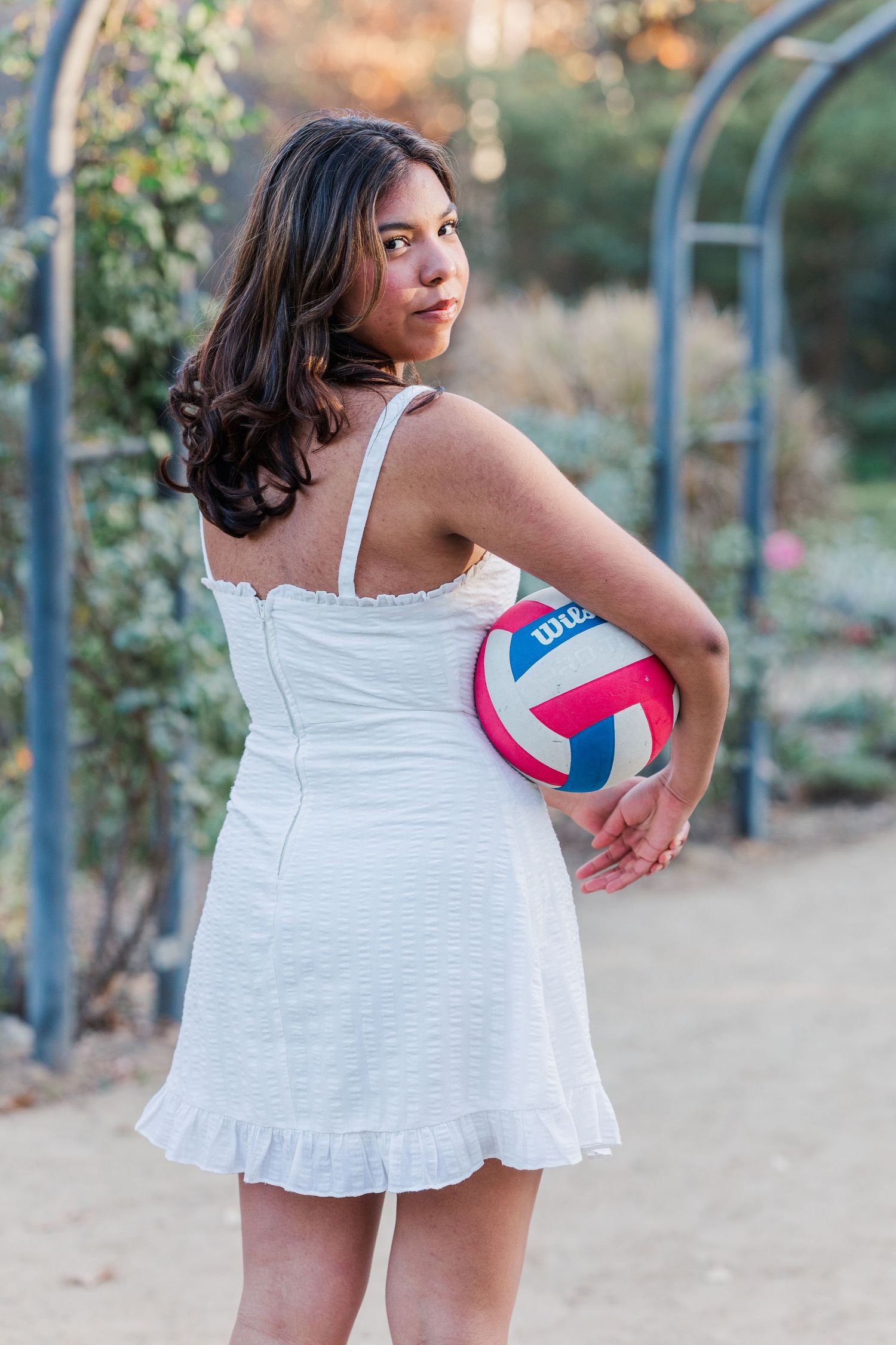 Athletic portrait in white dress holding colorful volleyball in garden setting.