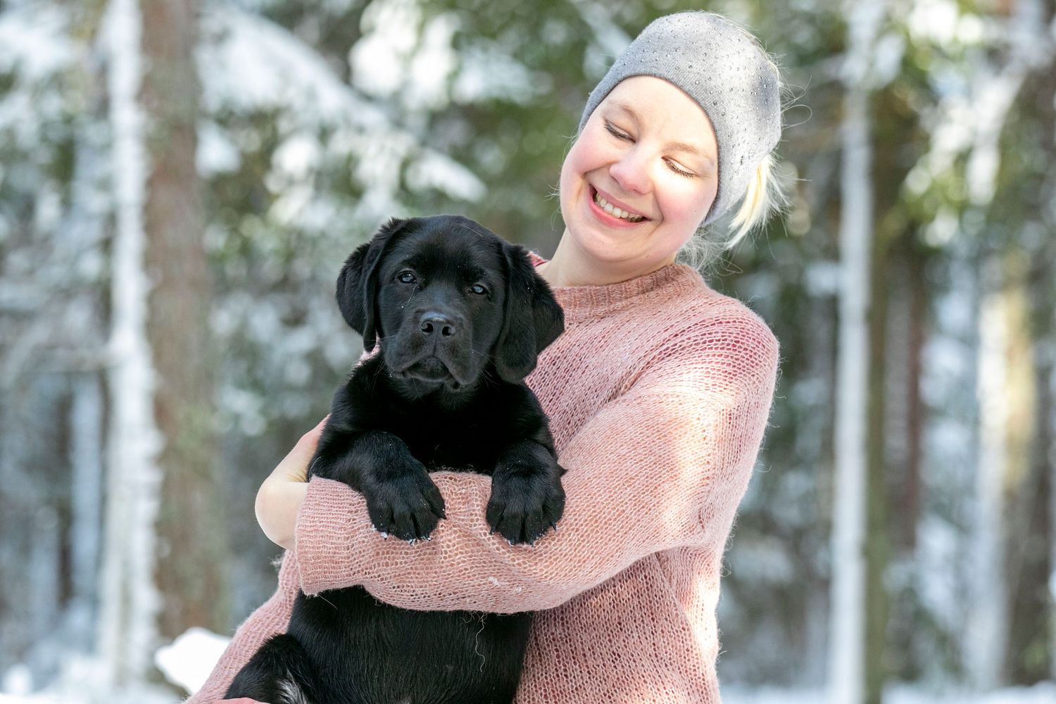 Person in pink sweater hugs black Labrador puppy in snowy winter forest.