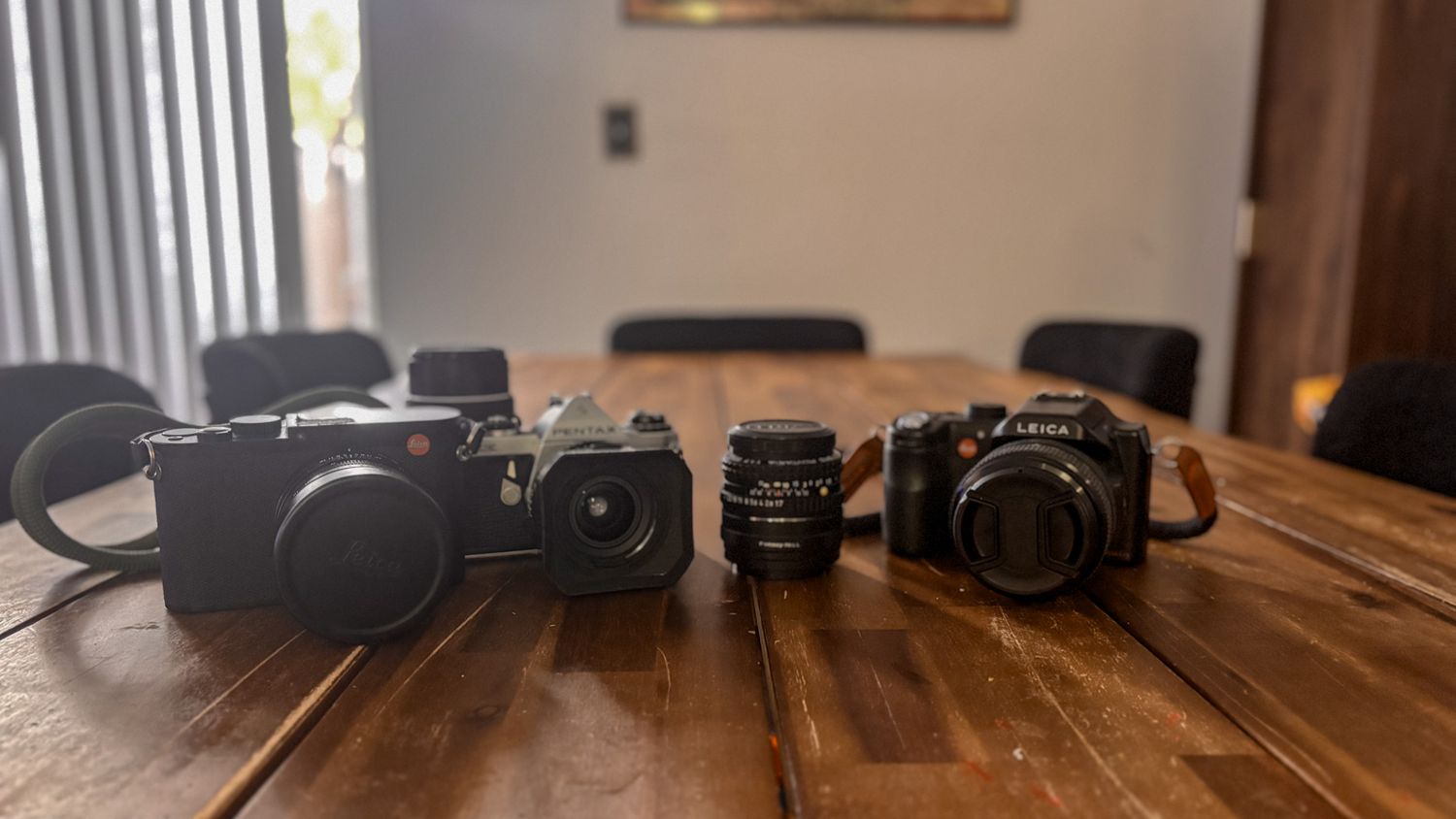 Multiple vintage cameras and lenses lined up on wooden dining table.