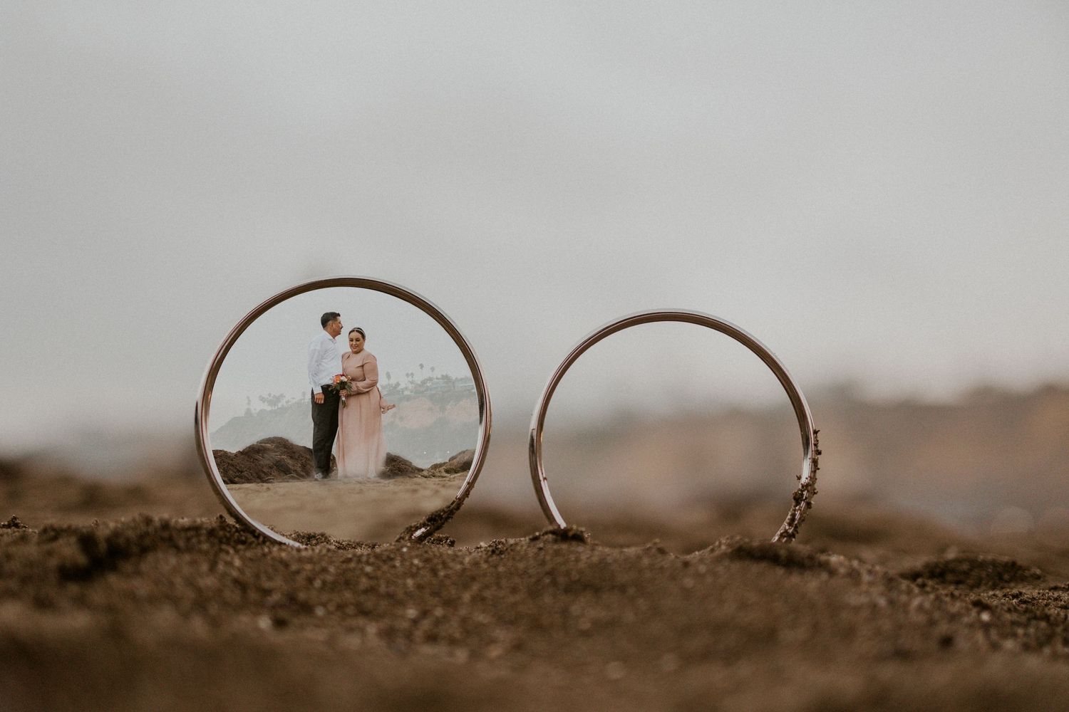 Creative wedding ring photo showing couple's reflection in one band while placed on sandy surface.