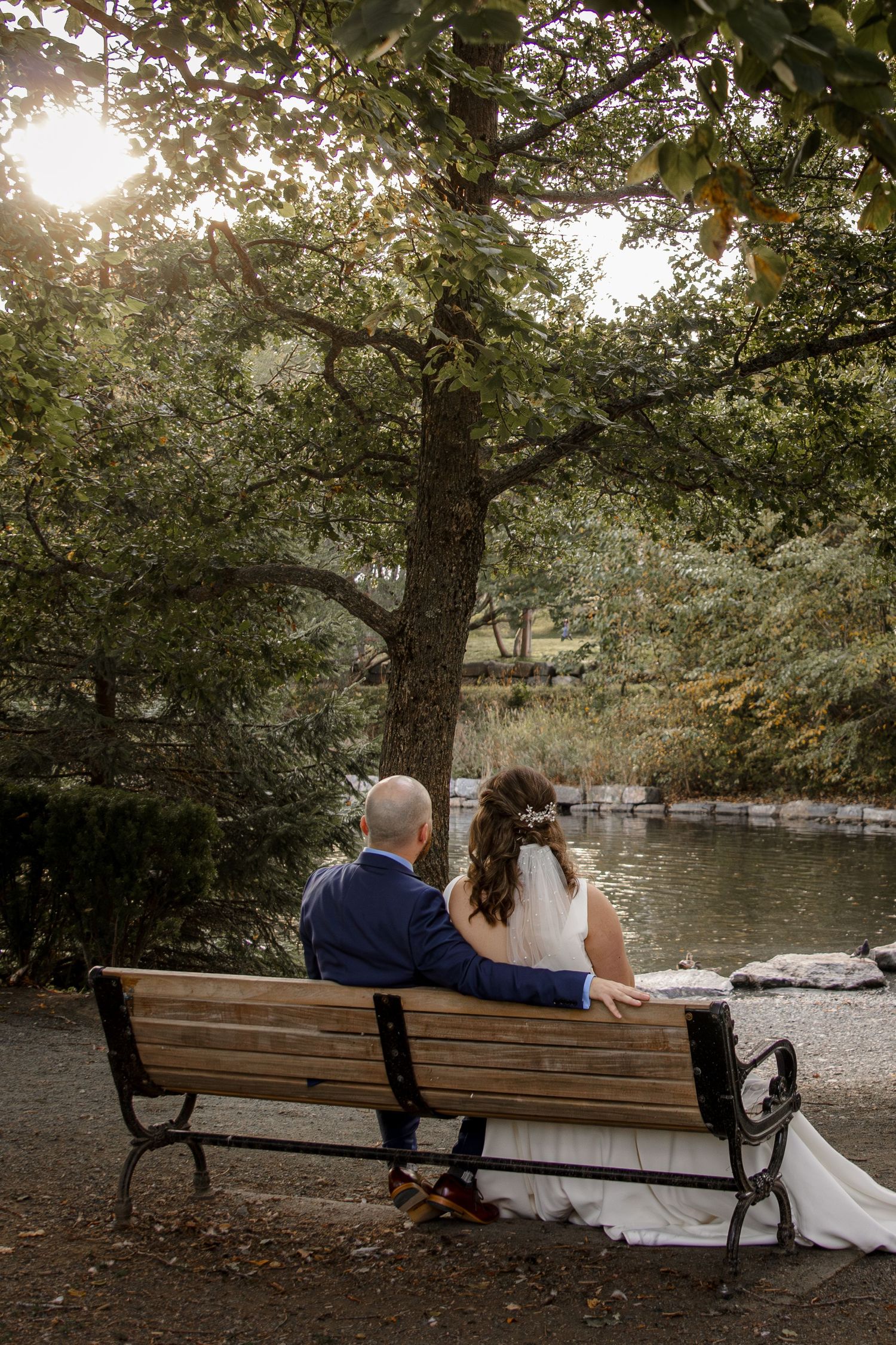 A couple sits together on a bench in bowring park by a tranquil pond at sunset while surrounded by leafy trees.