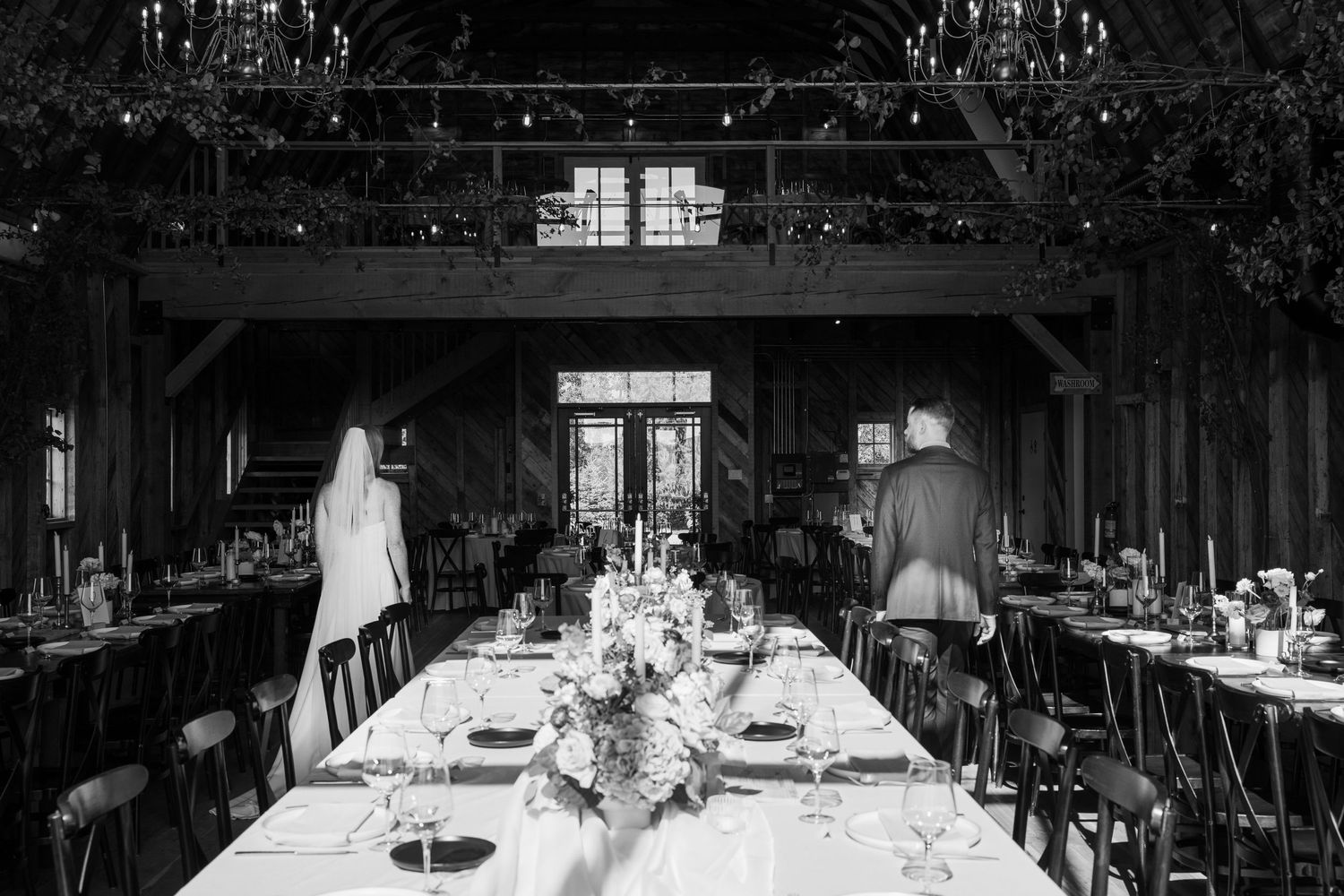 A black and white photo of an elegant wedding reception table setup in a rustic wooden barn venue.