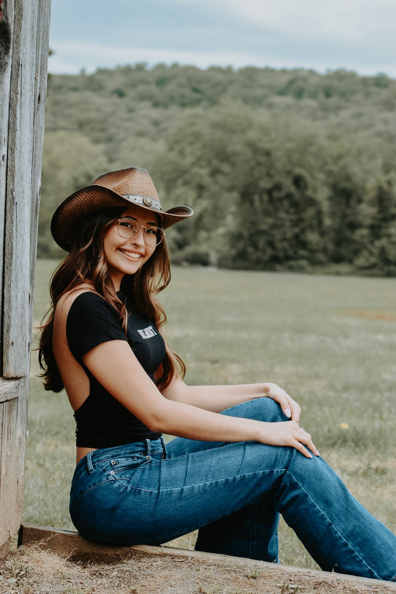 Casual portrait session in jeans and hat sitting against rustic wooden structure.
