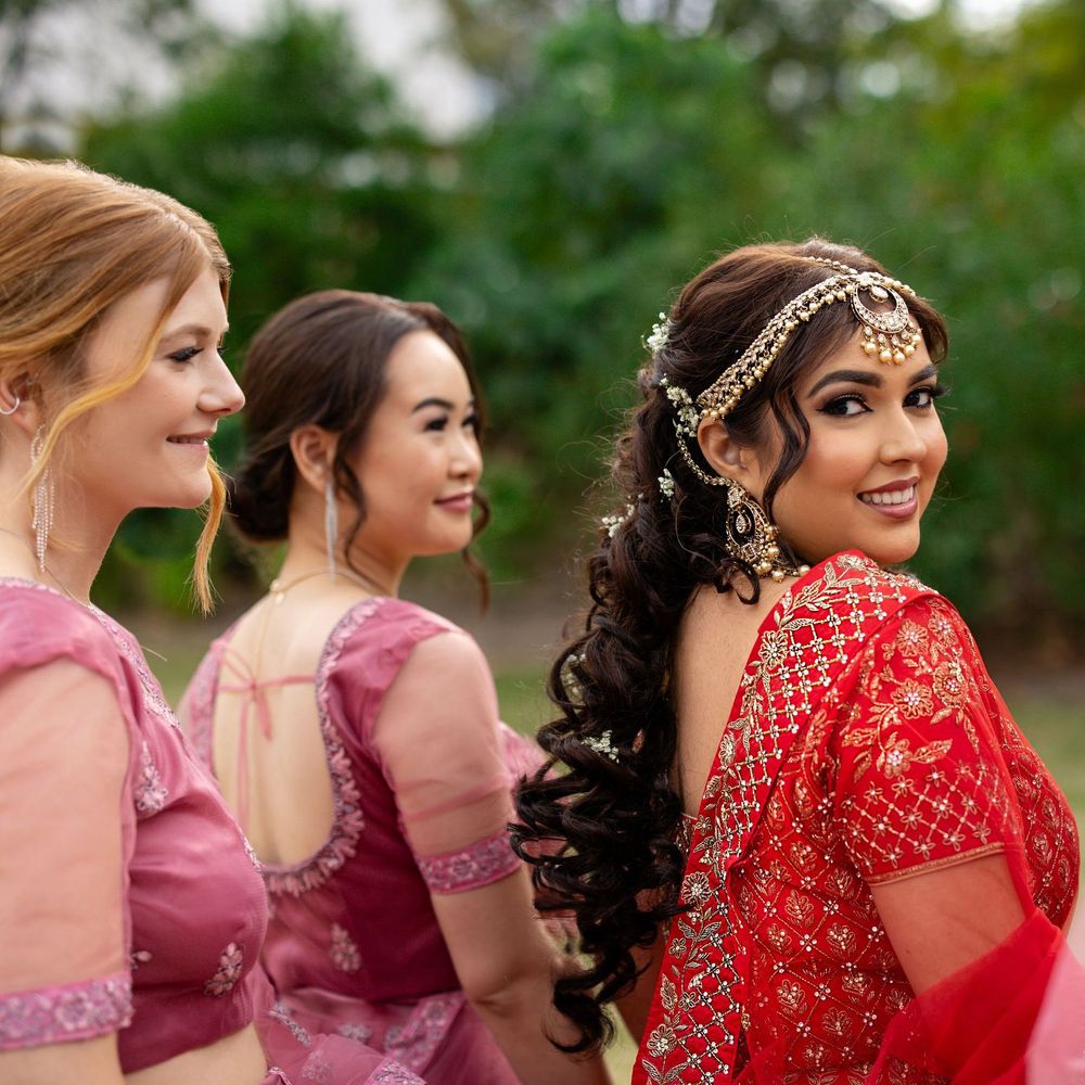 Indian bride in red lehenga smiling with bridesmaids in pink outfits outdoors.