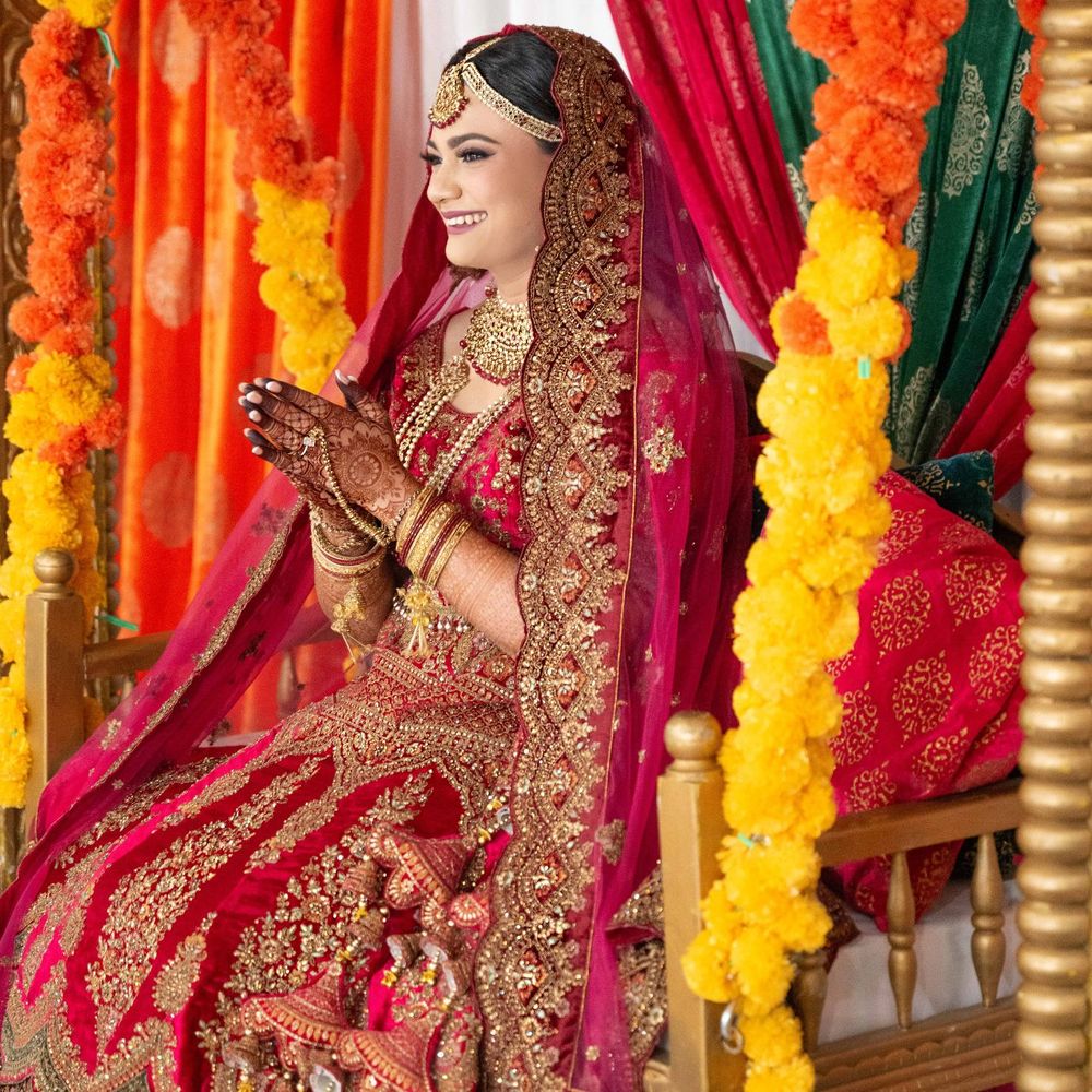 Radiant South Asian bride in red and gold lehenga seated at colorful floral wedding mandap.