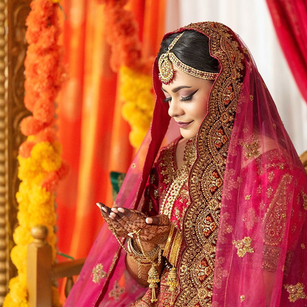 Beautiful South Asian bride in ornate red bridal lehenga holding ceremonial items at decorated mandap.