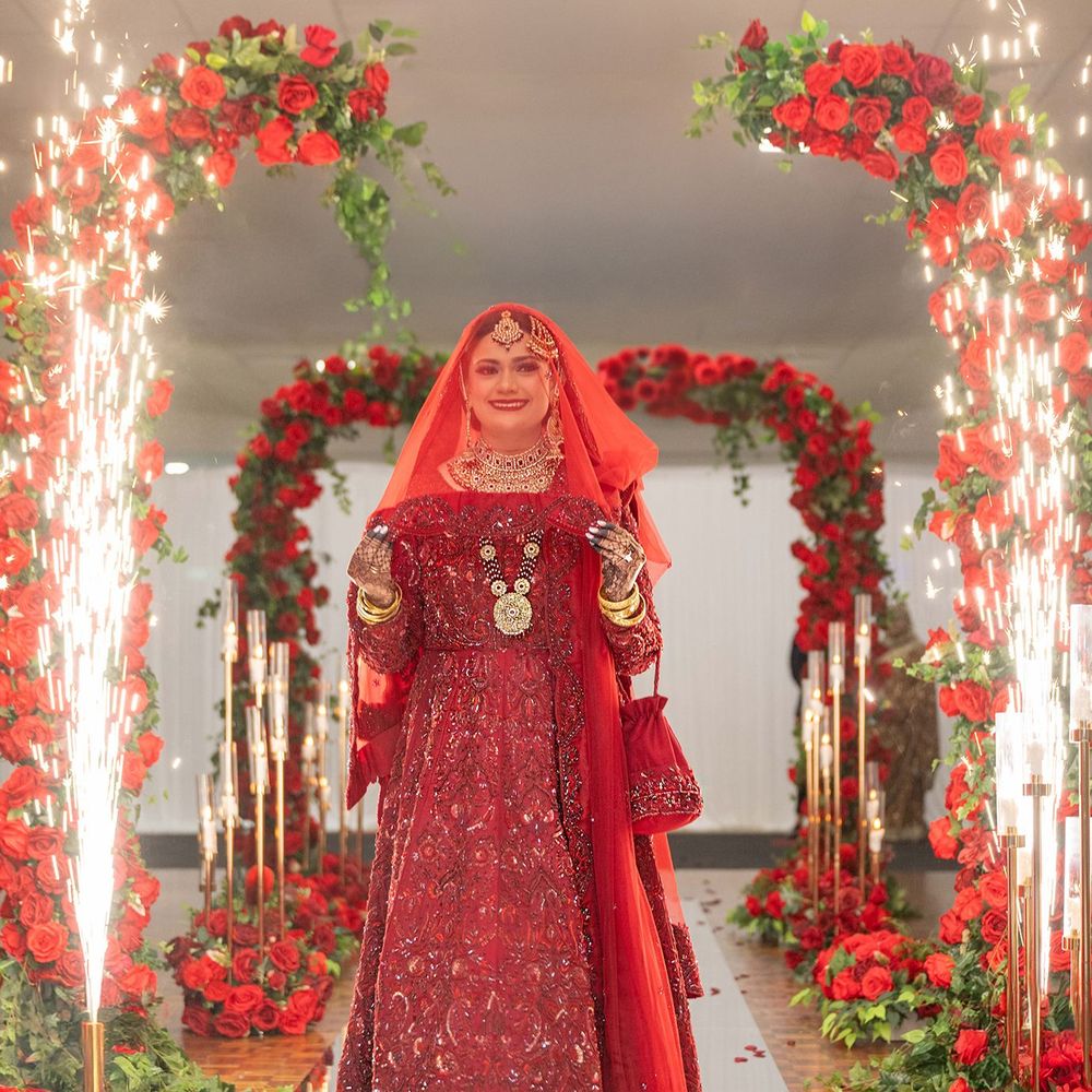 Indian bride in red bridal lehenga walking through sparkling fireworks and red floral arch.