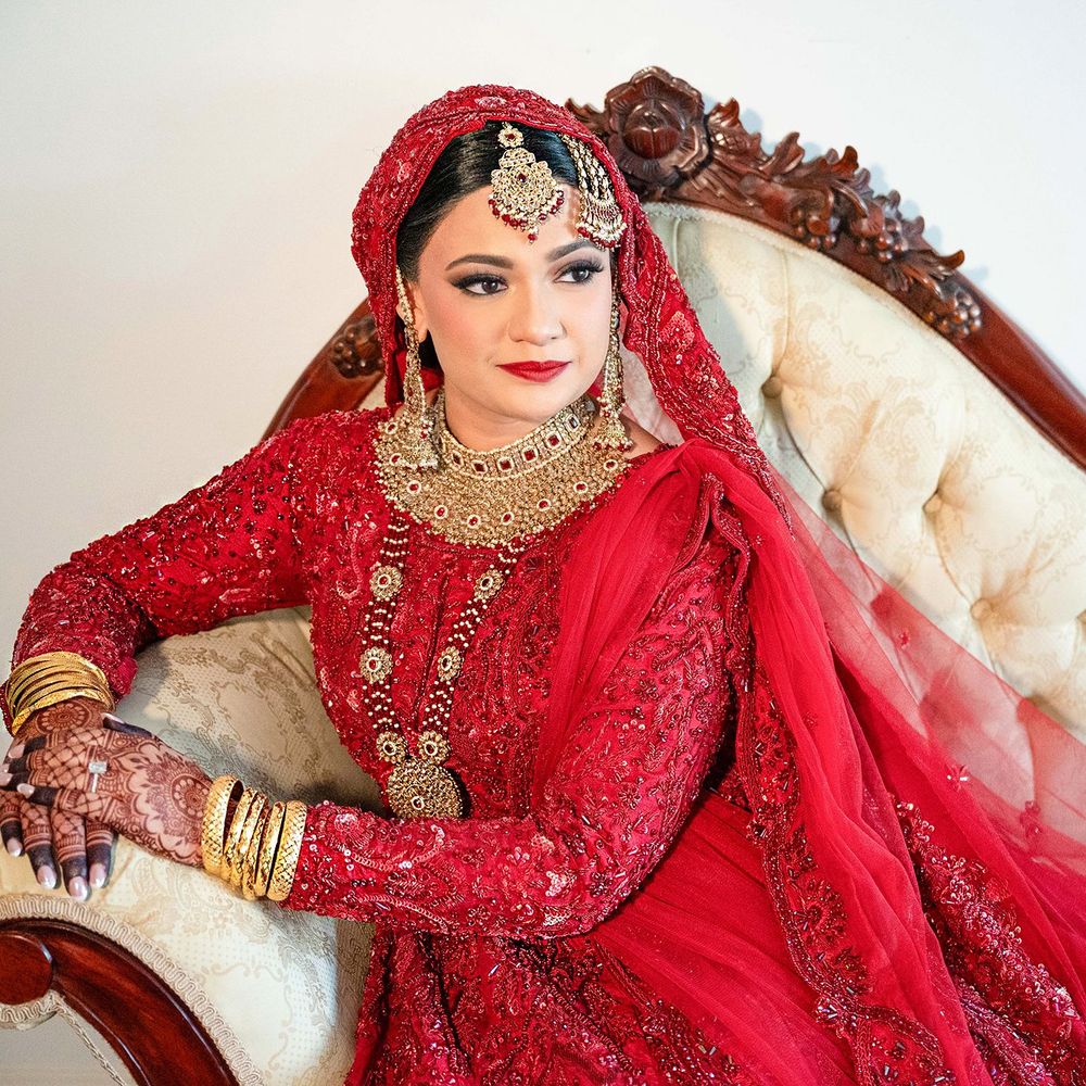 Indian bride in red embroidered bridal lehenga with gold jewelry seated on vintage sofa.