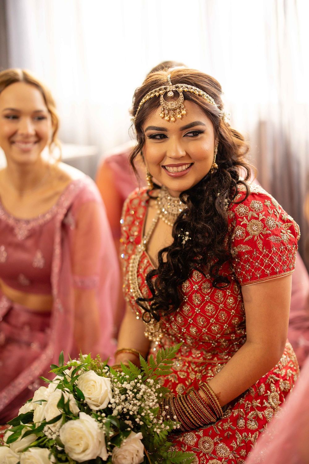 Smiling Indian bride in red lehenga with gold jewelry holding white roses bouquet.