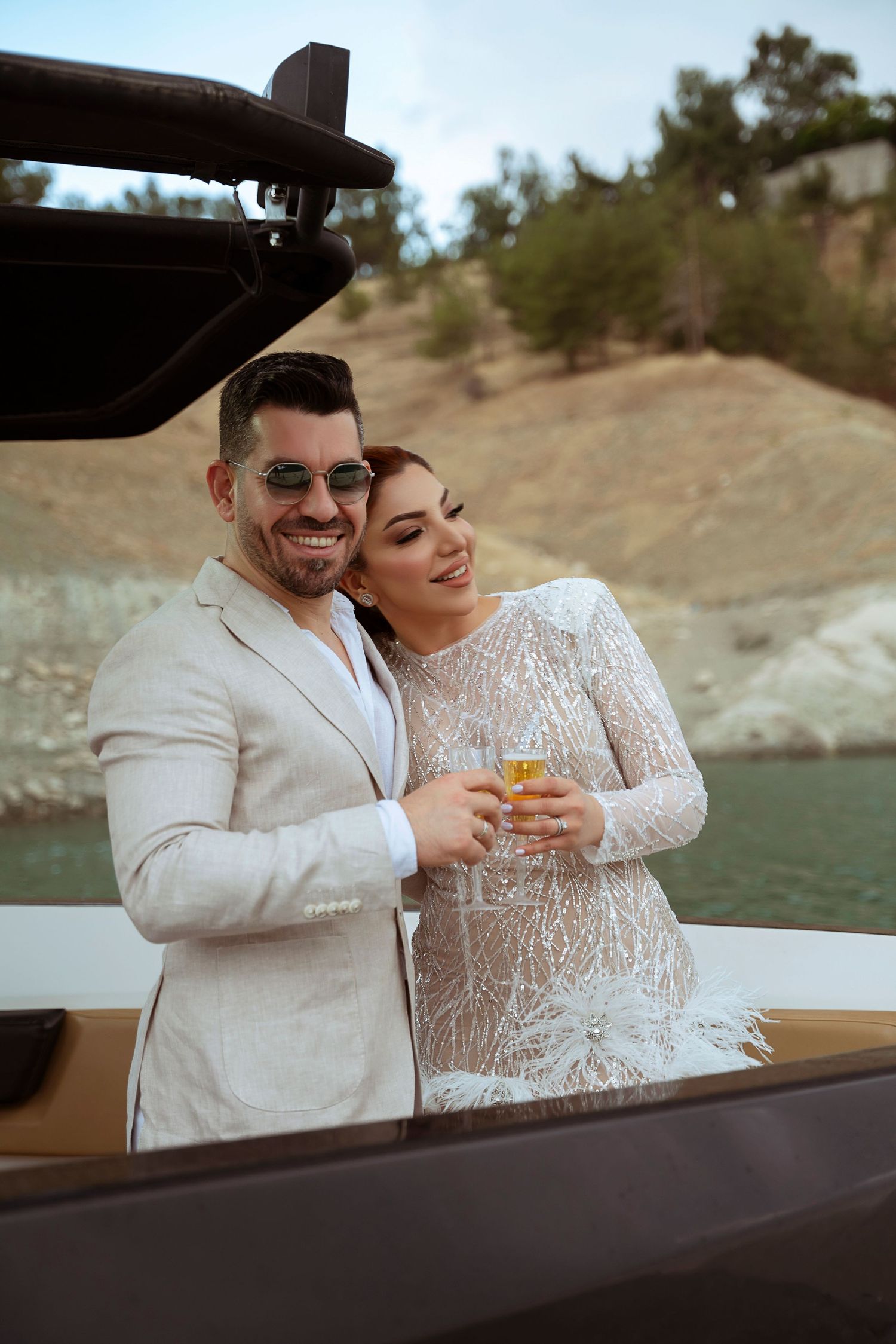 A happy couple in wedding attire share a drink while standing on a boat near a scenic lake and hillside.