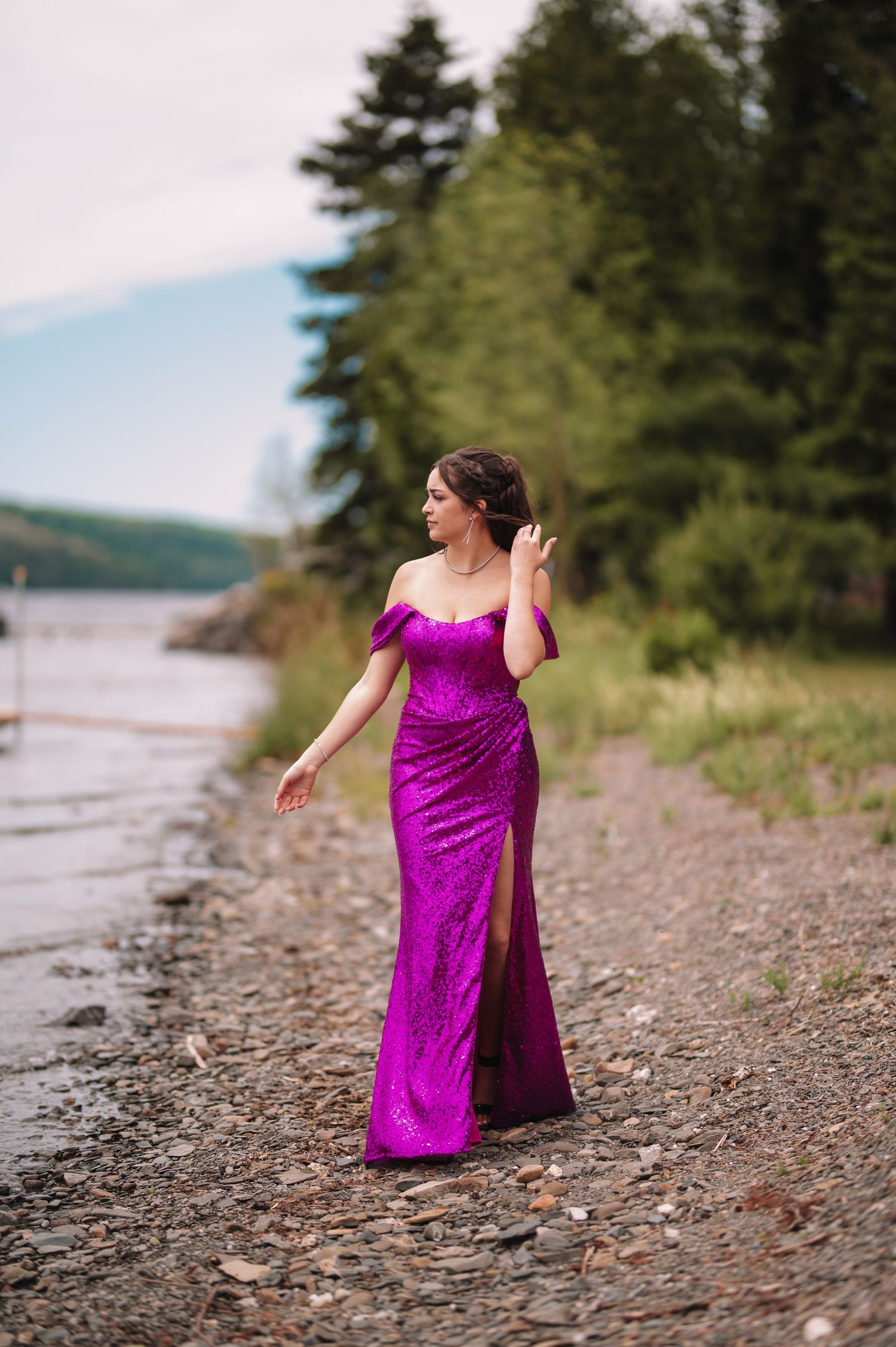 Woman in sparkling magenta gown poses elegantly on rocky beach shoreline with forest backdrop.