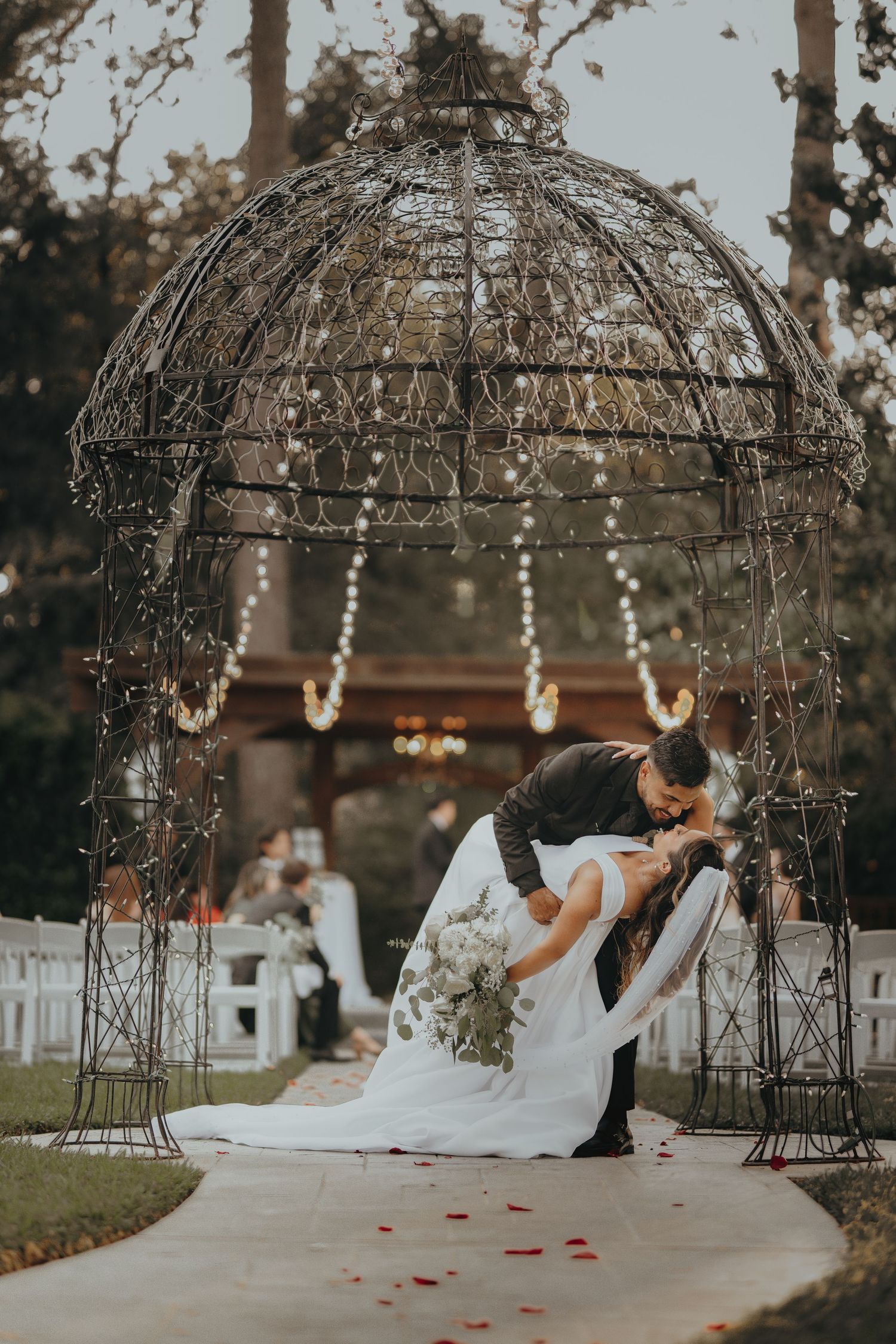 Wedding couple shares intimate moment under ornate metal gazebo decorated with lights and rose petals.