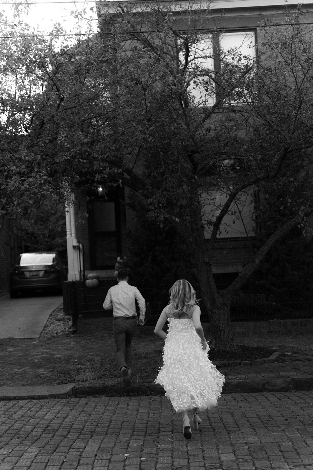 A couple runs playfully down a brick street in black and white photography.
