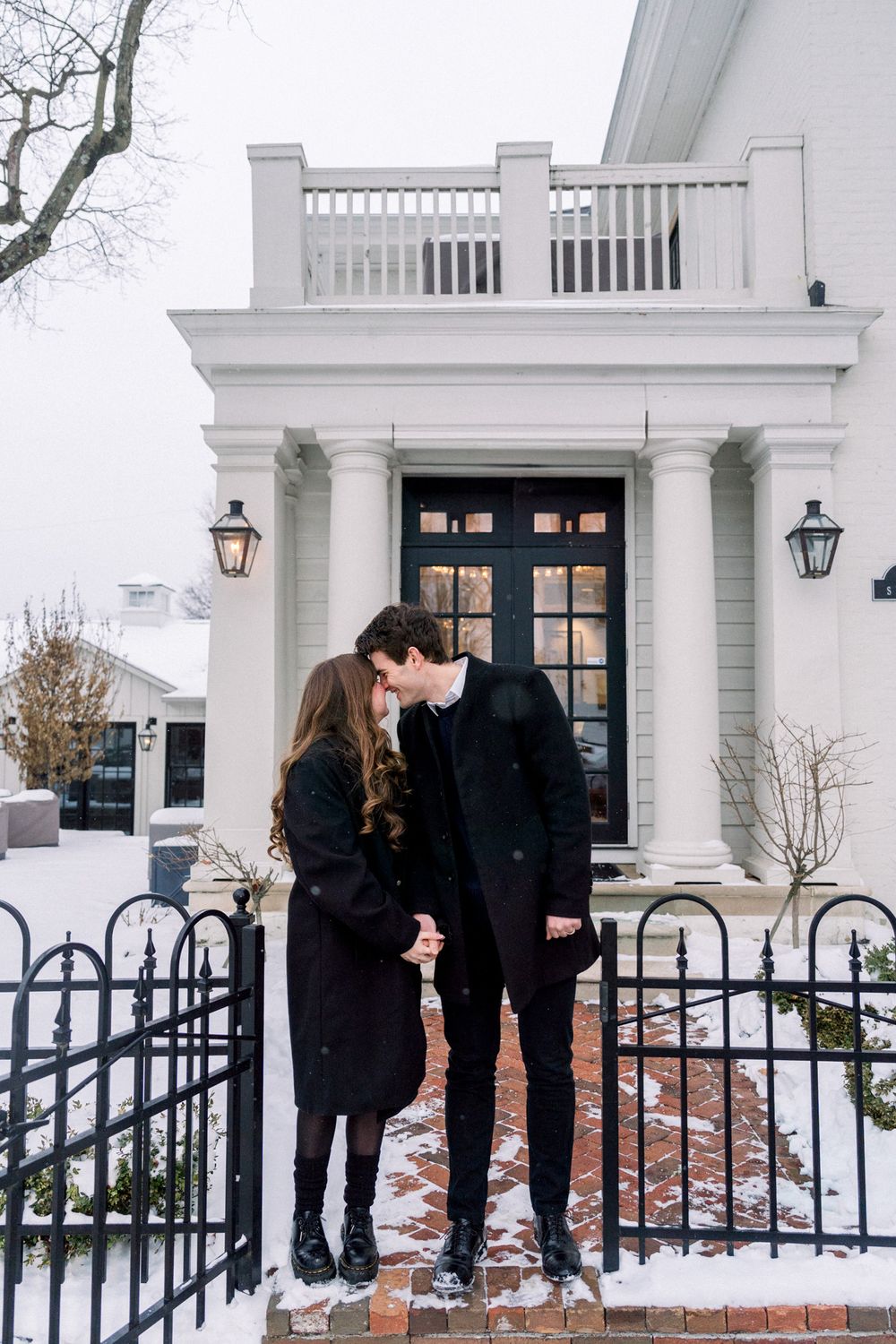 Couple in dark coats embracing on snowy steps of white colonial building.