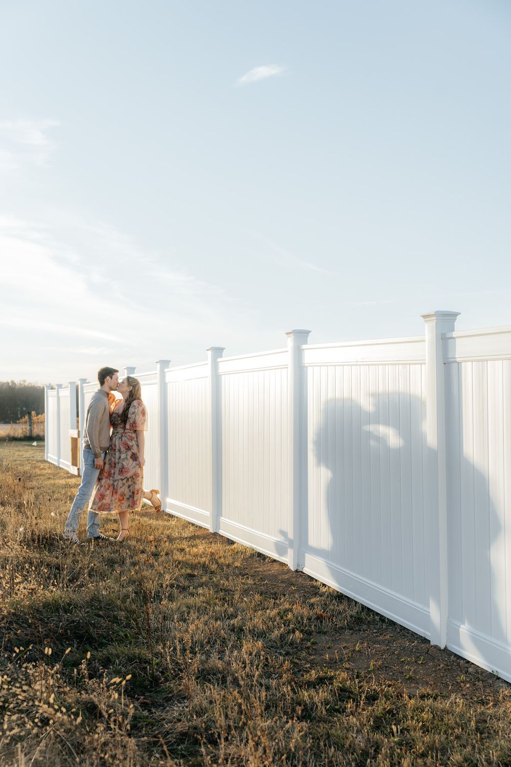 Couple poses against white fence creating dramatic shadow during sunset.
