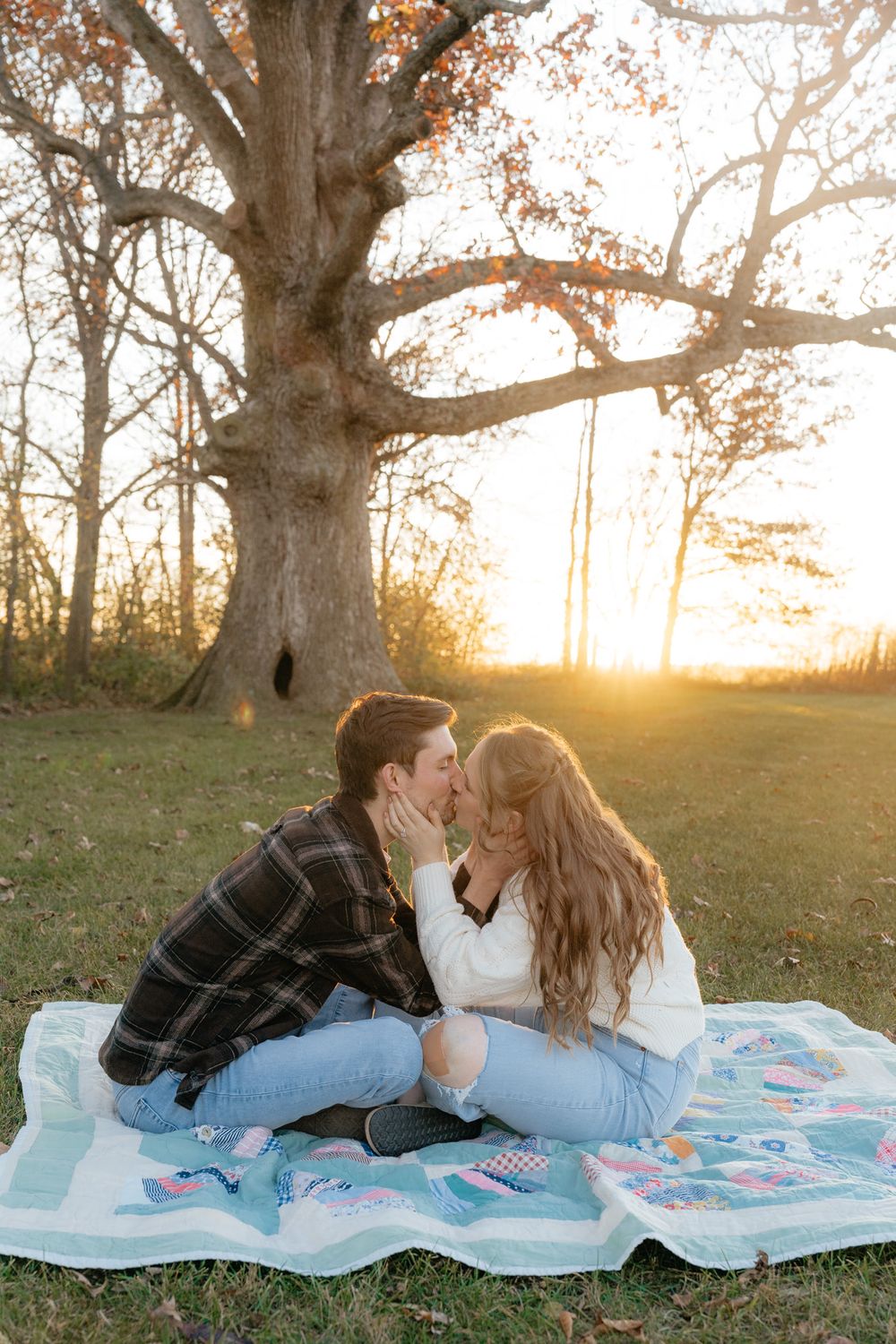 Romantic couple sharing an intimate moment on a picnic blanket at sunset under a large tree.