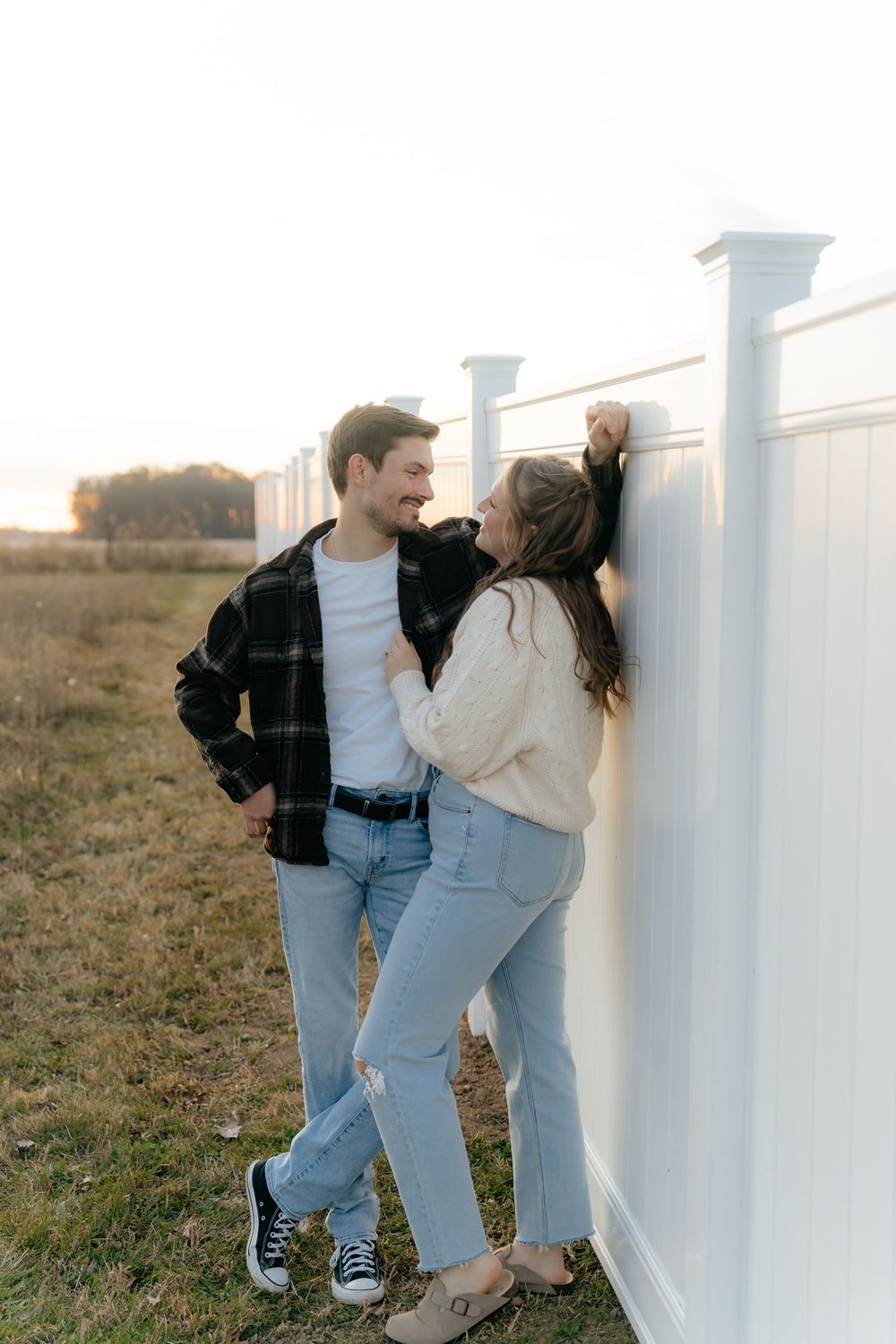Couple in casual wear sharing an intimate moment against a white fence at sunset.