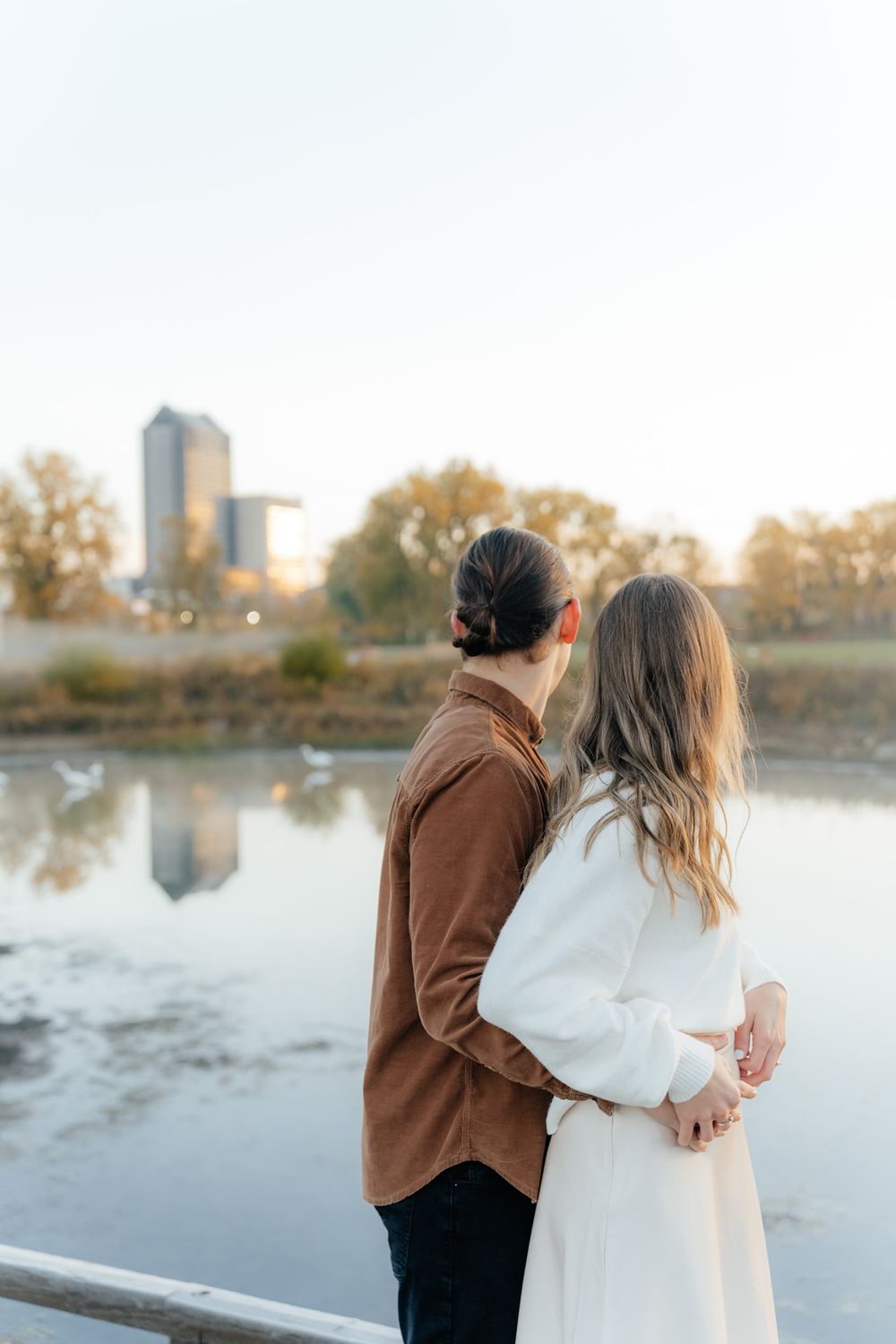 Couple in brown and white outfits standing by a reflective lake with city buildings in the distance.