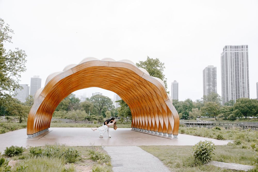 Couple dancing under a wooden arch sculpture in a park with city skyline backdrop.