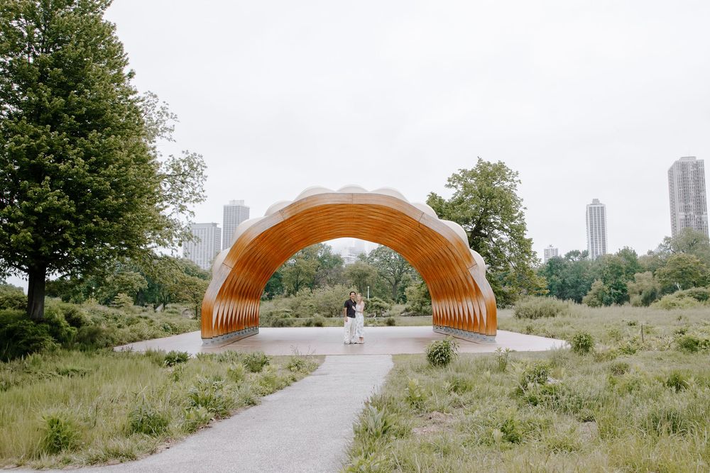 A wooden arch sculpture in a city park with a person standing beneath it, surrounded by greenery and skyline.