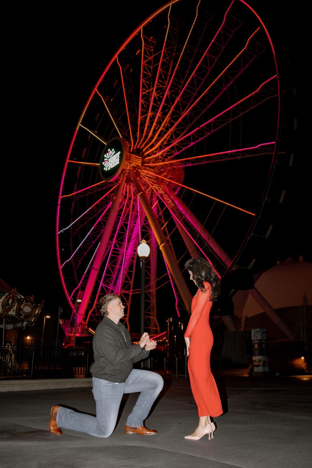 Man proposes on one knee to woman in red dress at night with illuminated Ferris wheel backdrop.