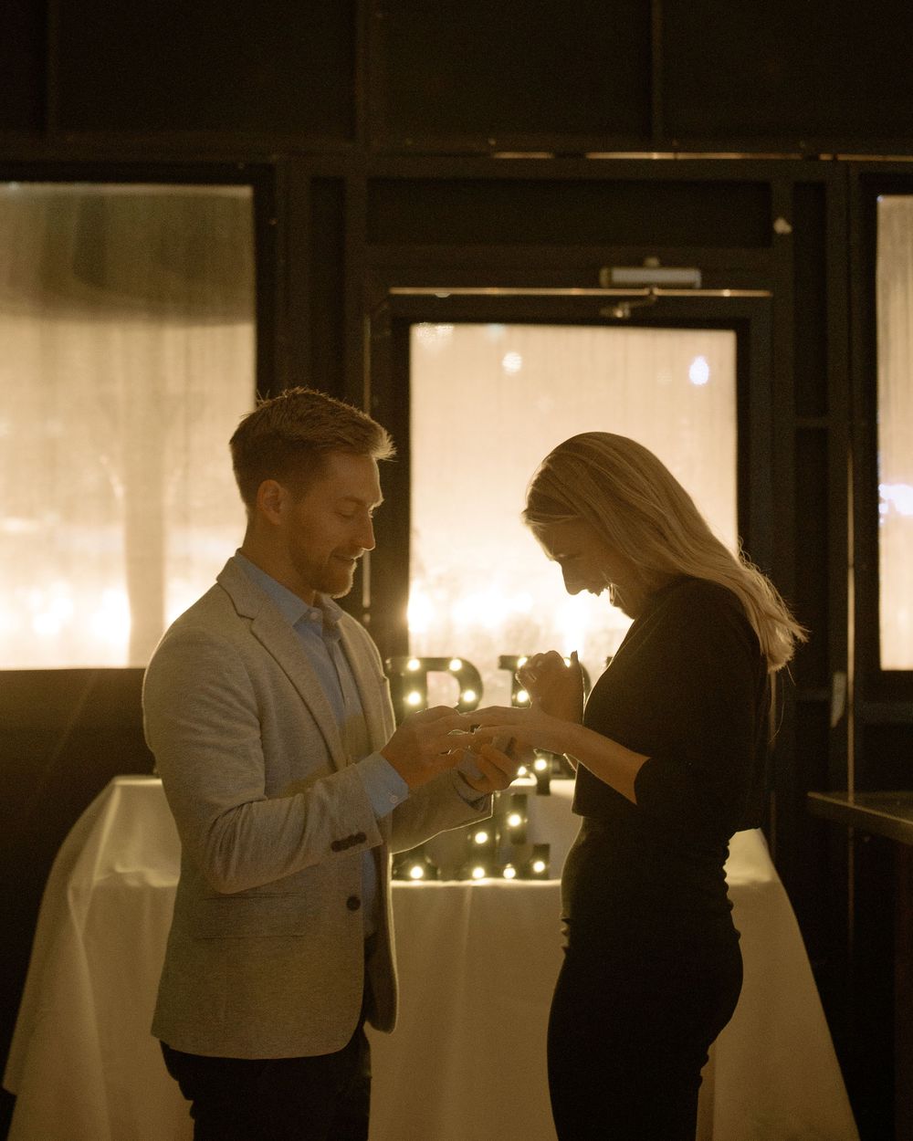 A couple shares a romantic moment in a candlelit restaurant, man in blazer presenting a ring to a woman in black dress.