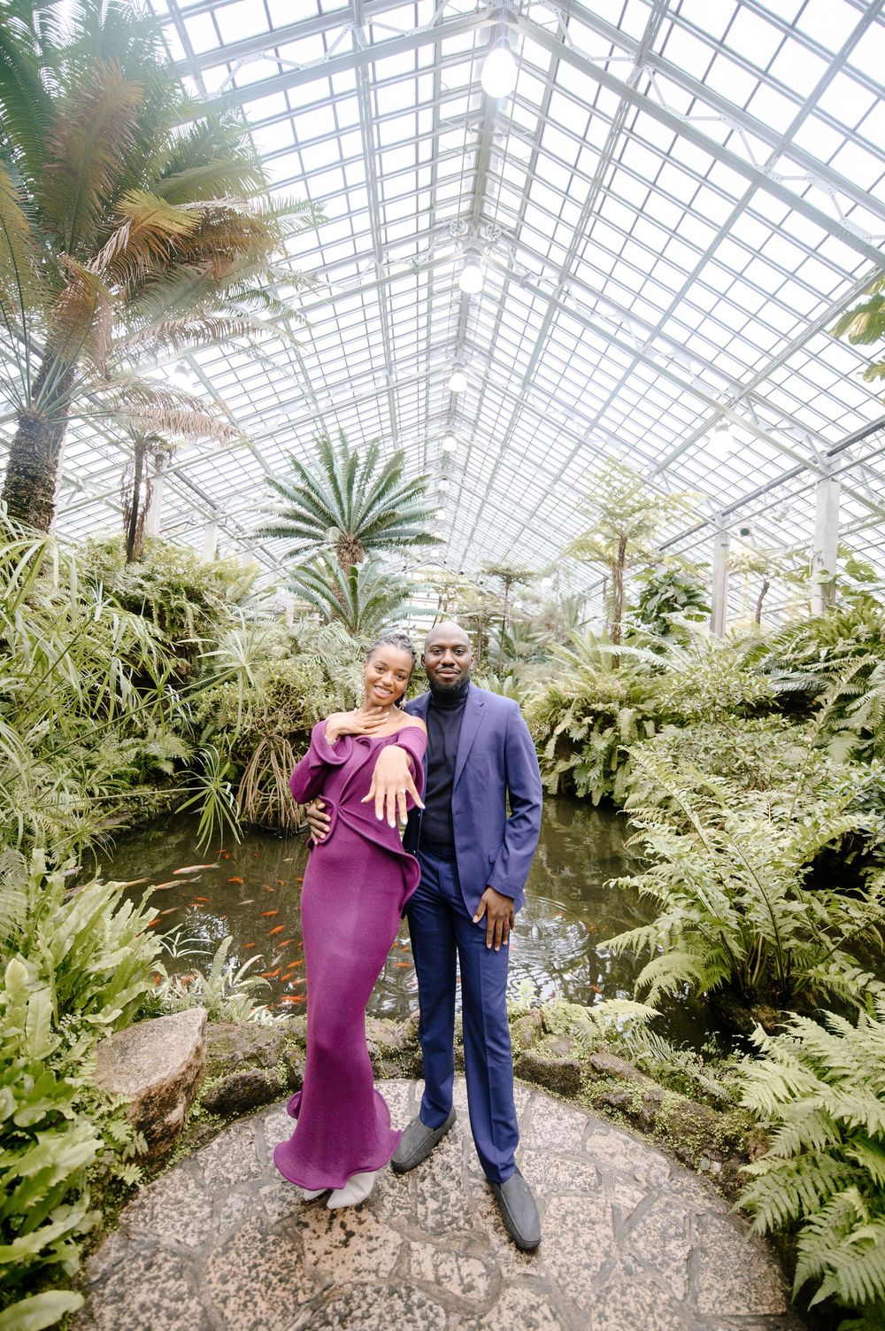 Couple poses in elegant attire inside a lush greenhouse botanical garden with tropical plants and glass ceiling.
