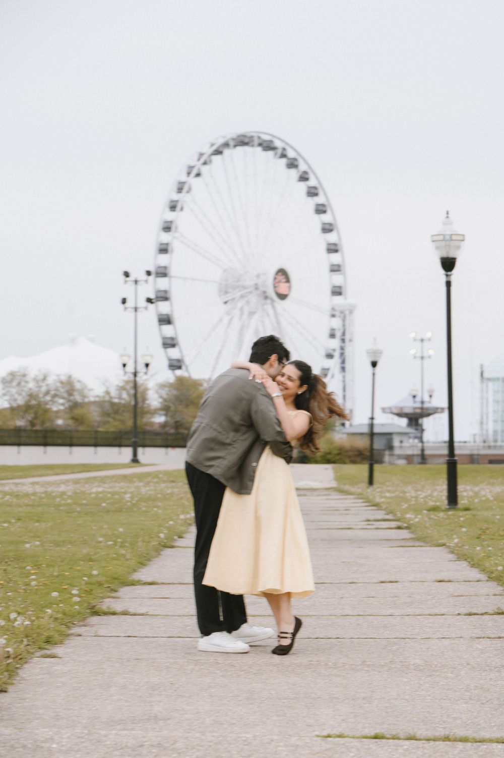 Couple embracing on a pathway with a large Ferris wheel in the background on a cloudy day.