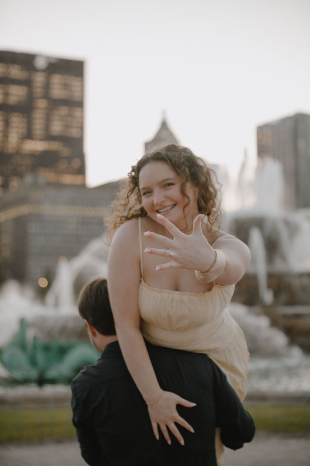 A joyful woman shows off her engagement ring while being carried by her partner near Buckingham Fountain in Chicago.