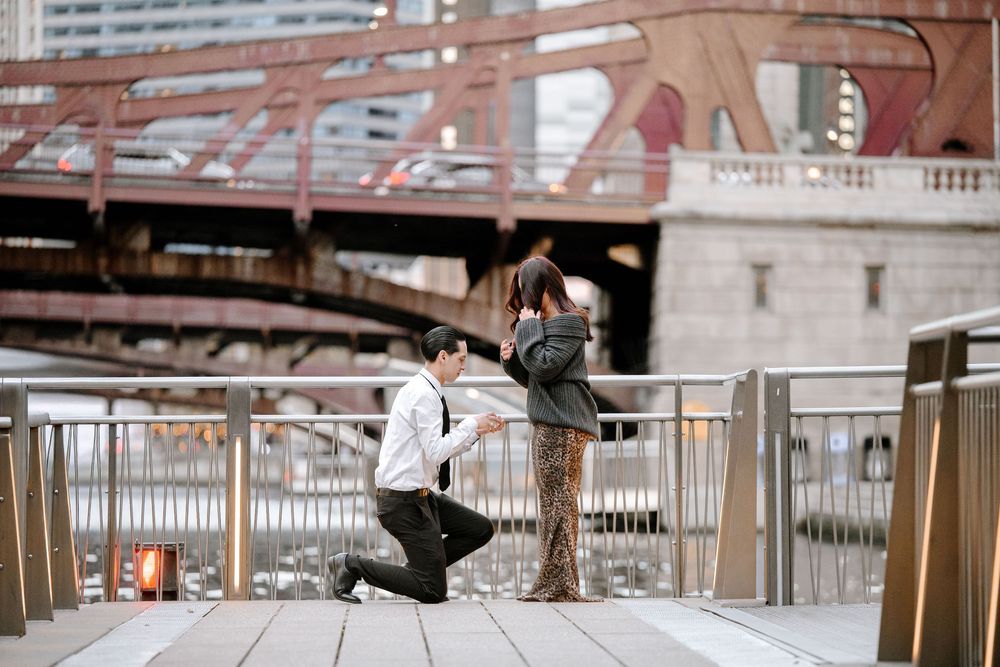 Man proposes to woman on one knee on a riverside walkway near a bridge in an urban city setting.