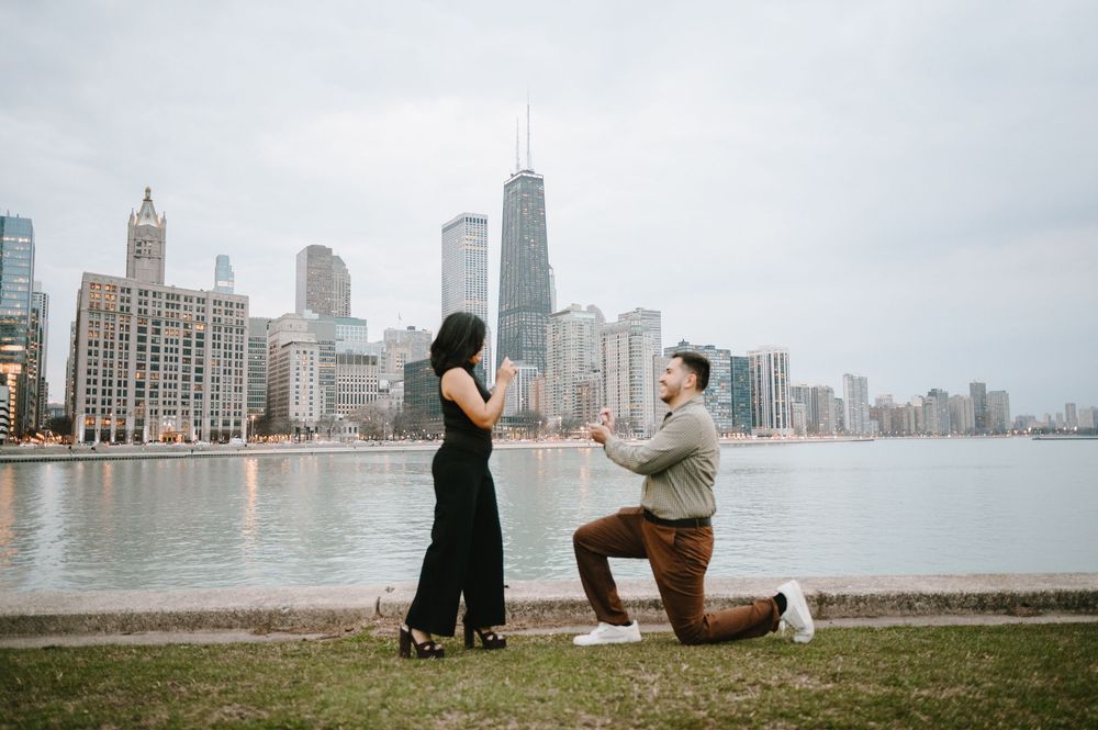 Man proposes to woman on one knee by Chicago lakefront with city skyline backdrop.