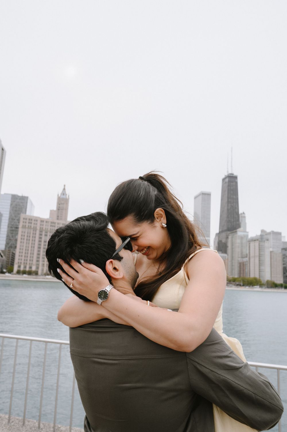 Couple embracing and smiling near Chicago waterfront with city skyline including John Hancock Tower in background.