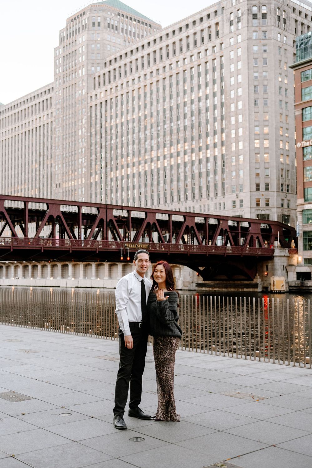 Couple posing on Chicago Riverwalk with historic iron bridge and large city buildings in the background.