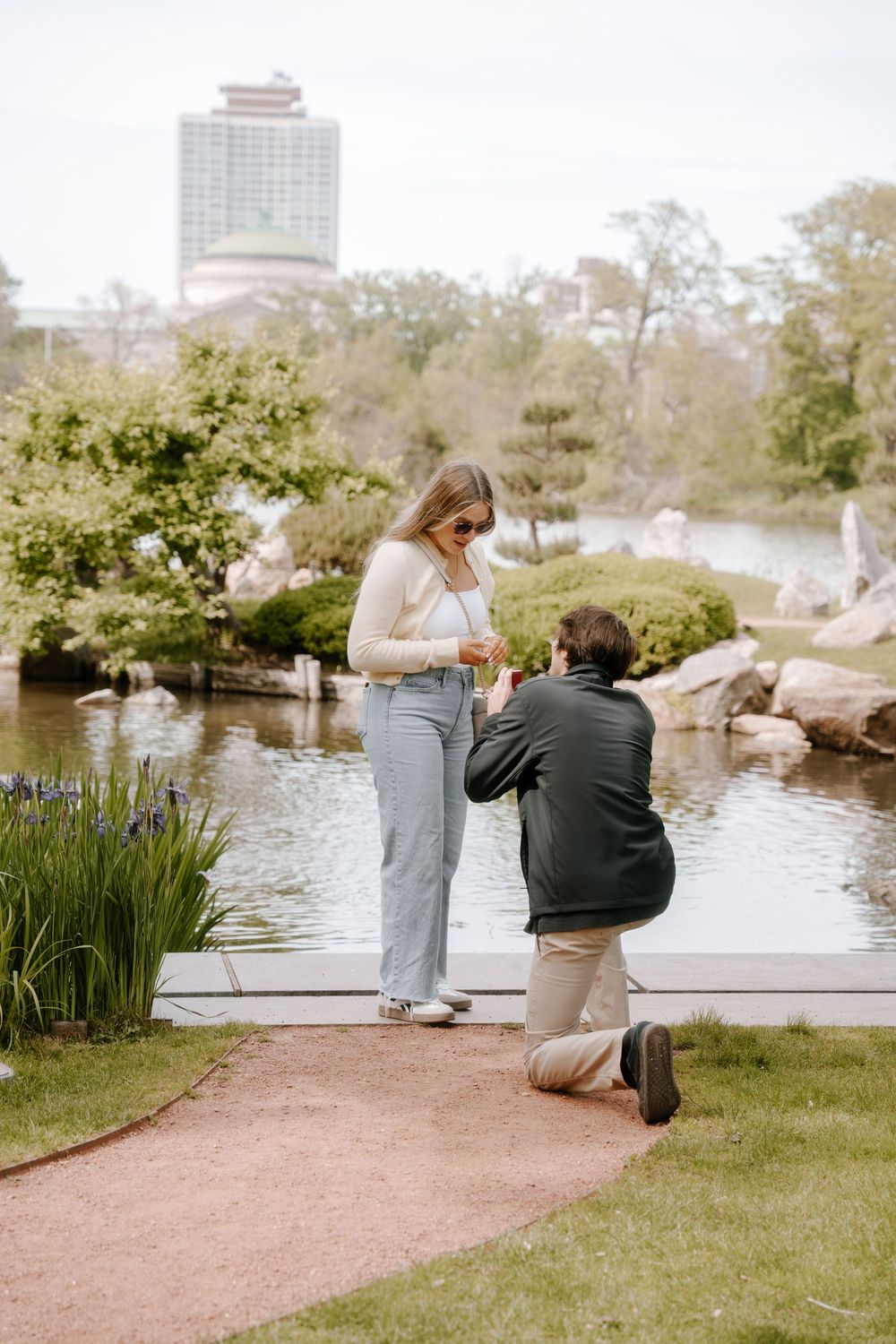 Man kneeling on one knee proposing to woman in a lush garden park with a pond and city skyline backdrop.