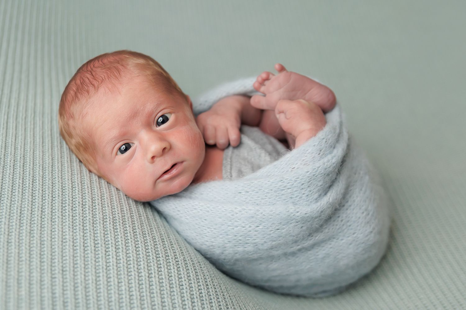 A newborn baby with a reddish tint to their hair, wrapped in a light blue blanket, lying on a textured pale green surface, looking at the ca