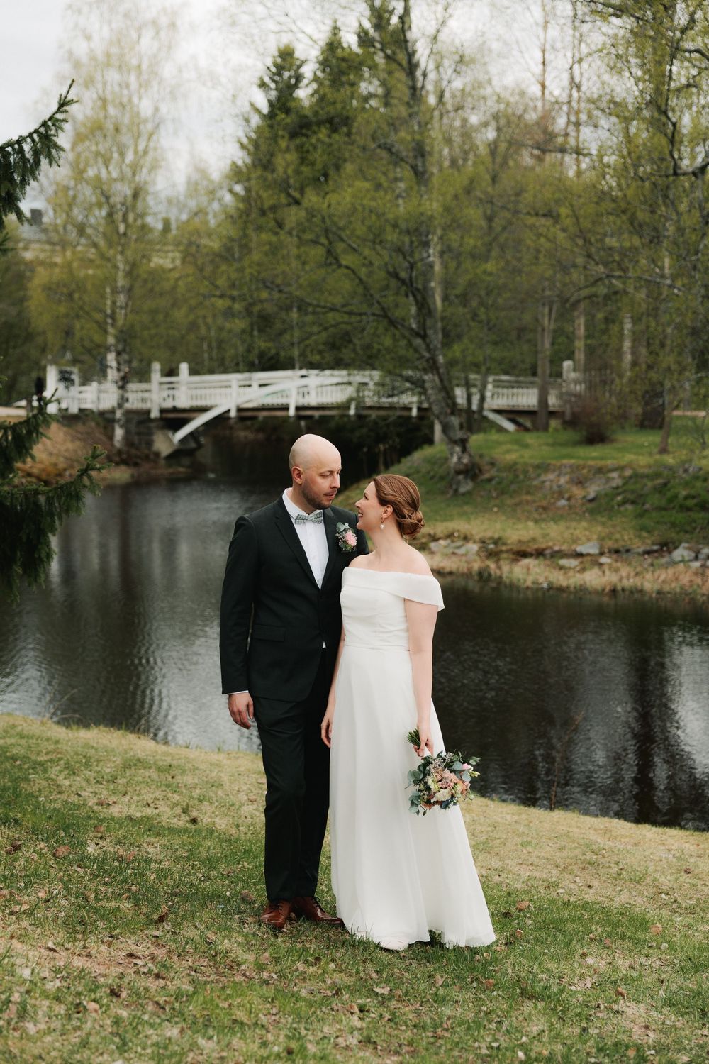 A couple in formal wedding attire stands by a serene pond with trees and a bridge in the background.