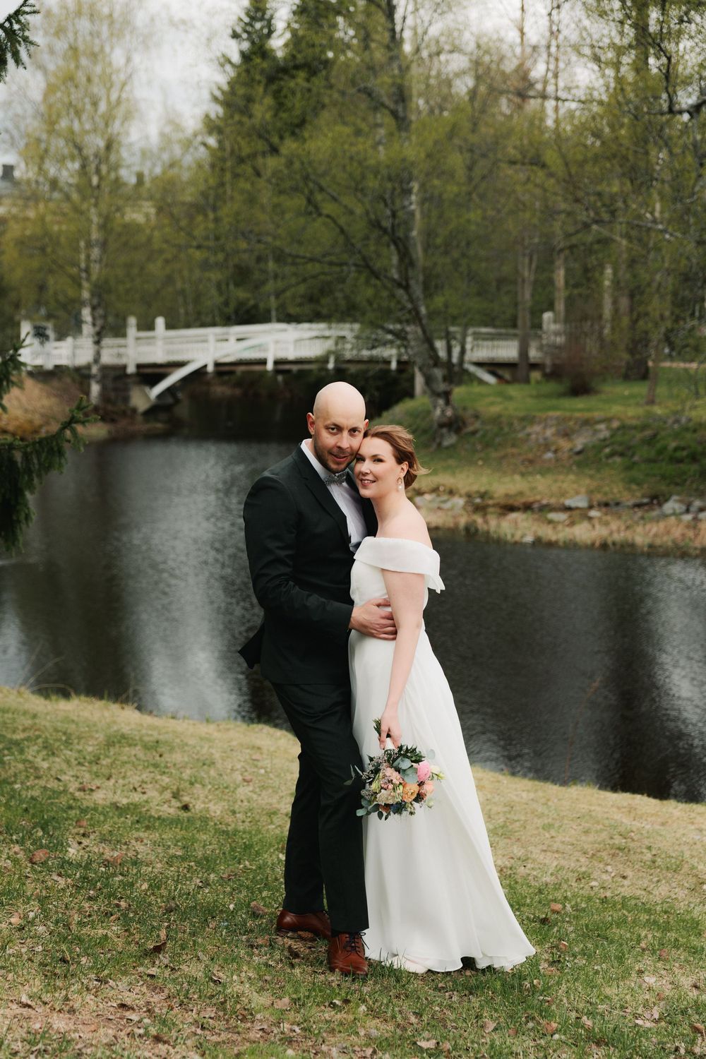 A couple in wedding attire embraces by a pond with a white bridge and spring trees in the background.